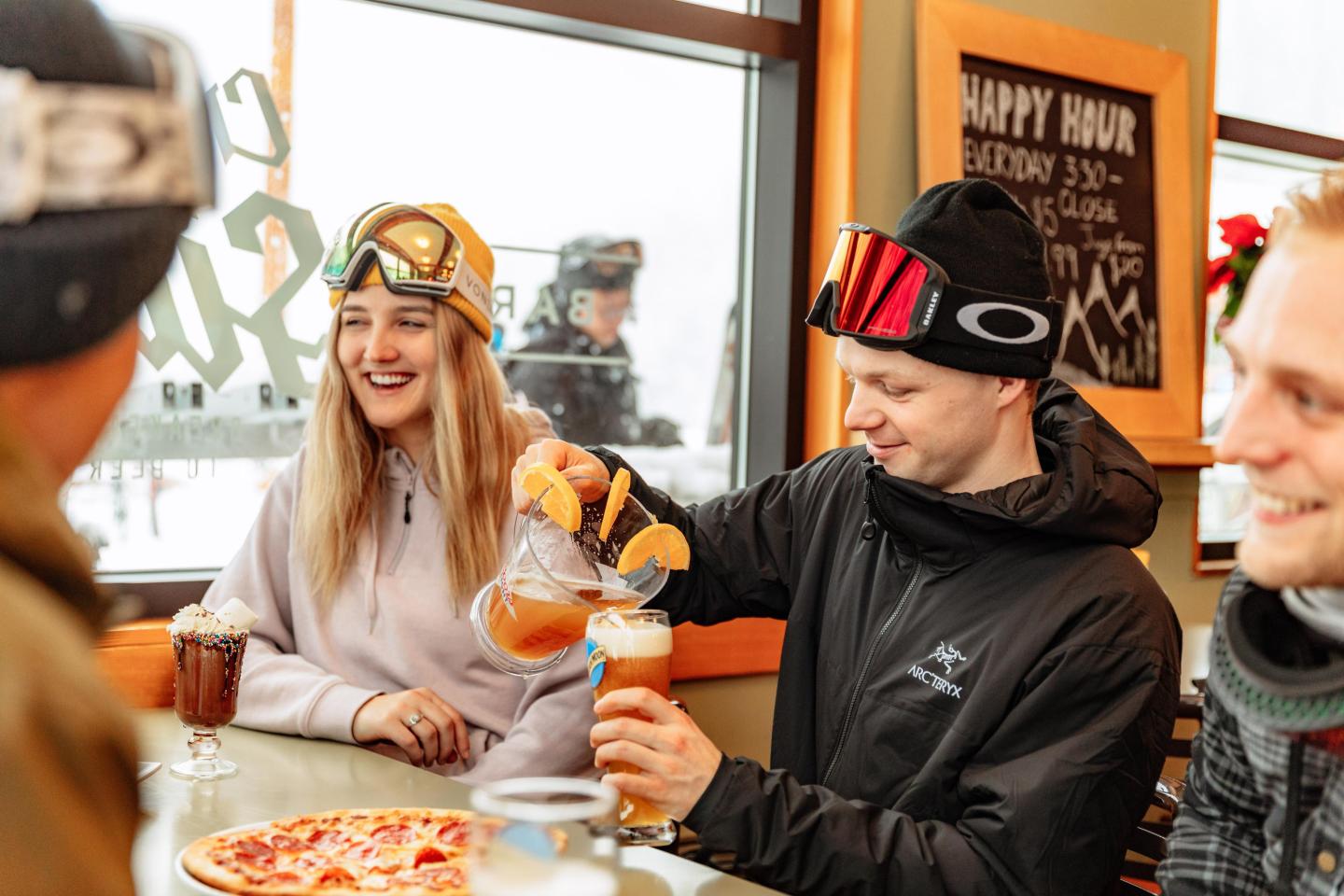 Group of friends in ski gear enjoying drinks and pizza indoors.