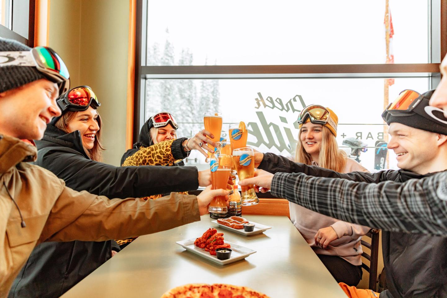 Group of friends in ski gear toasting with drinks in a cozy restaurant.