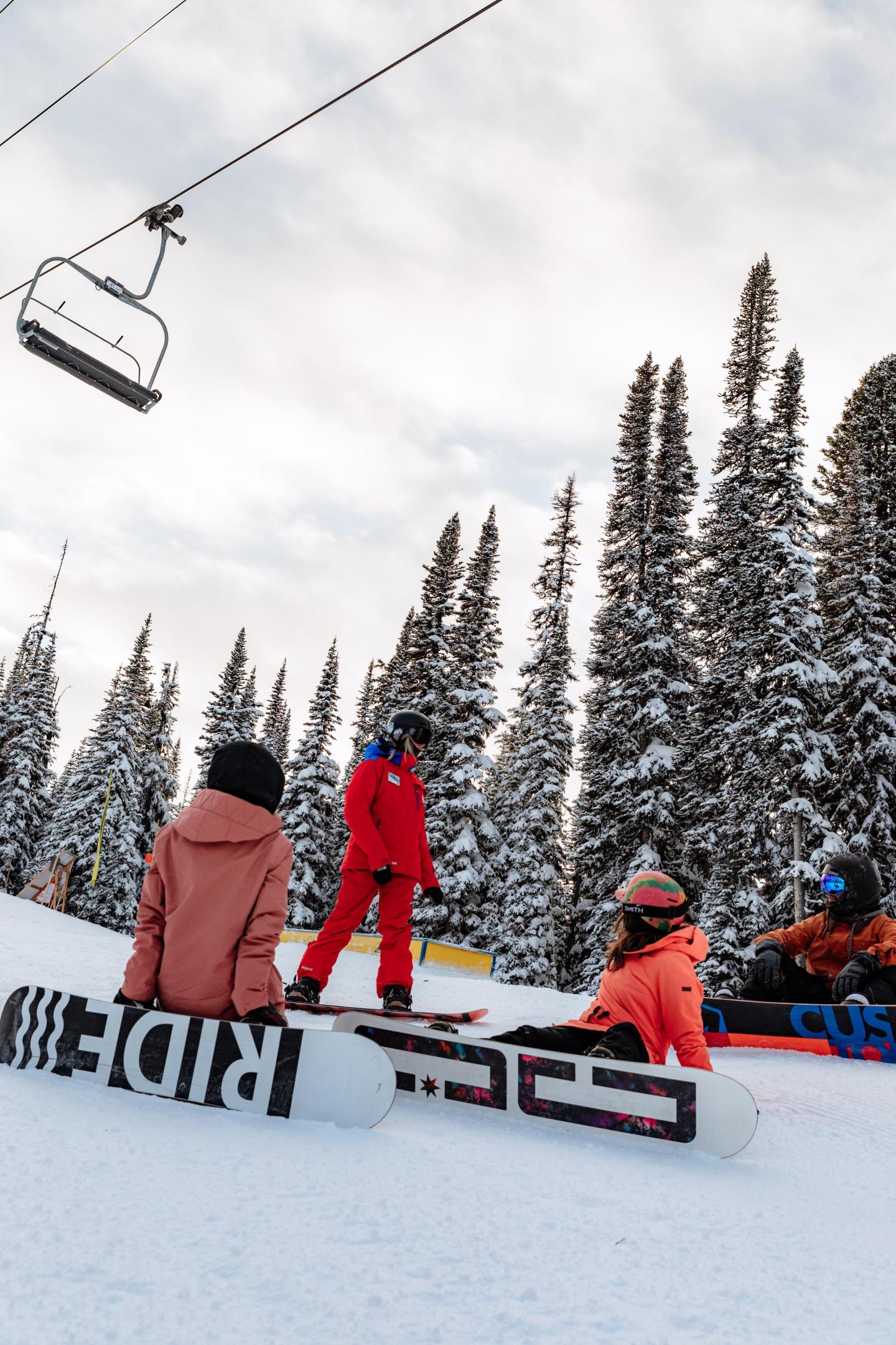 Skiers in colorful gear rest on a snowy slope beneath a cloudy sky.