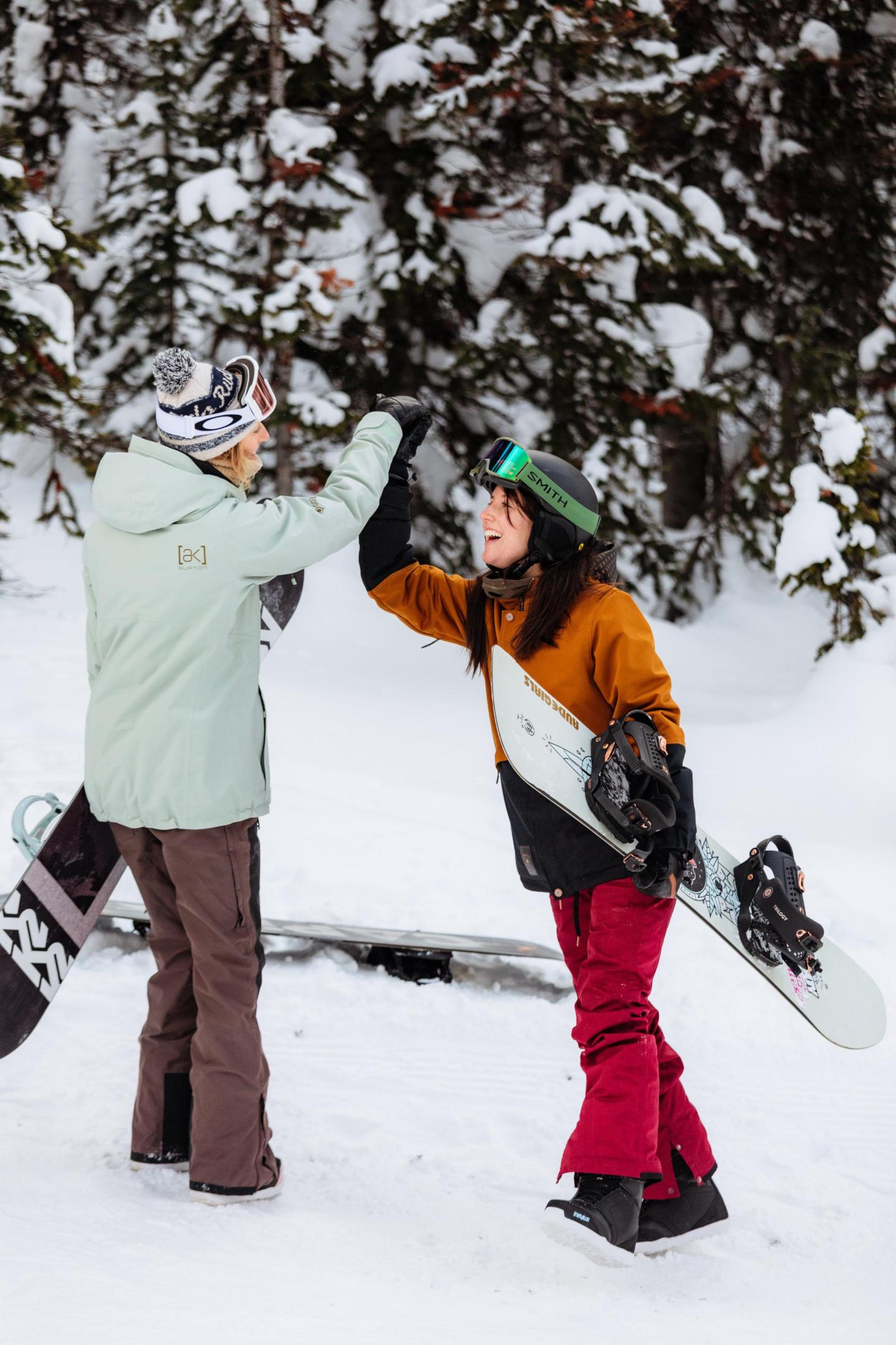 Two snowboarders high-five, surrounded by snowy trees.