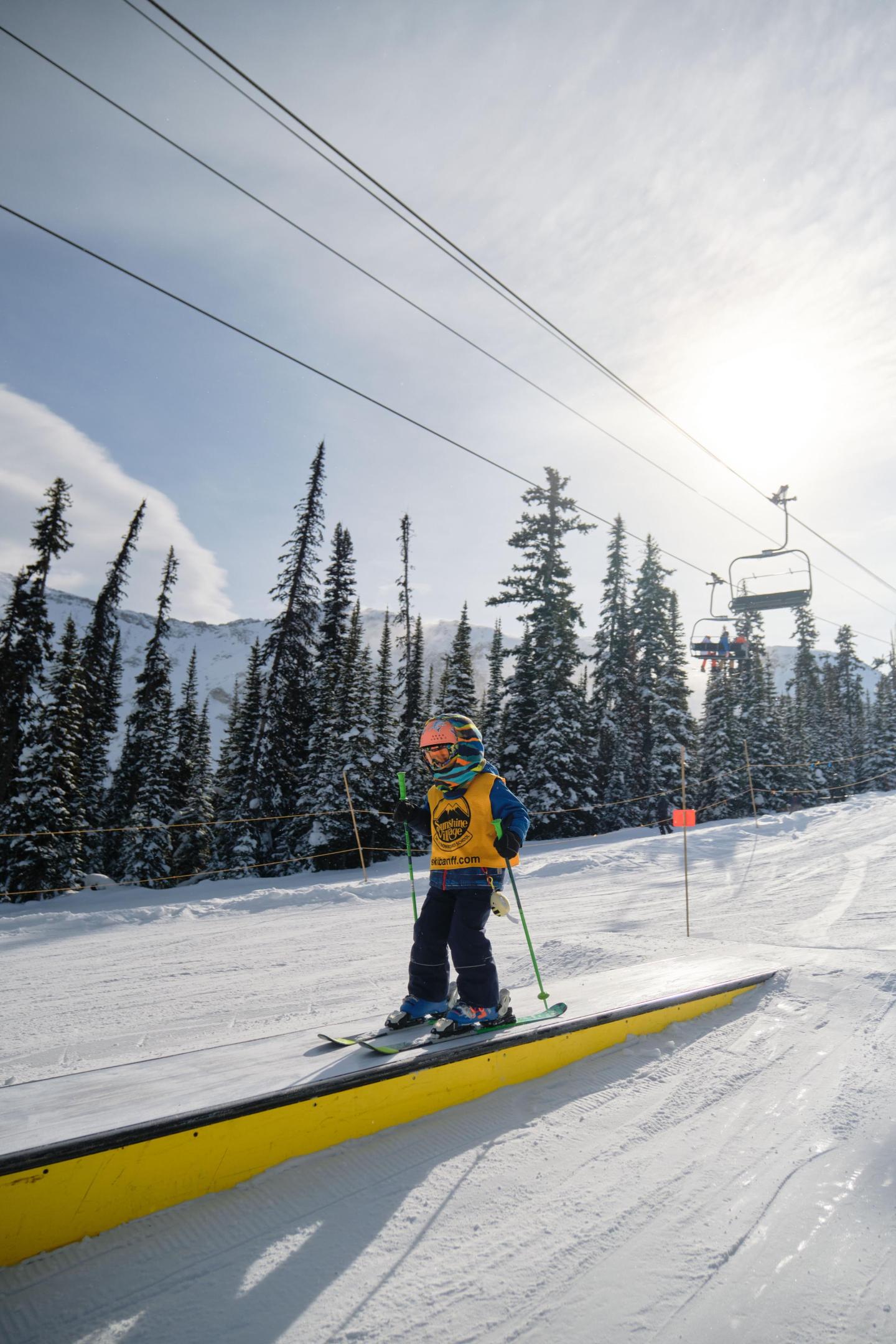 Skier in a yellow vest on a rail, snowy trees and ski lift in background.