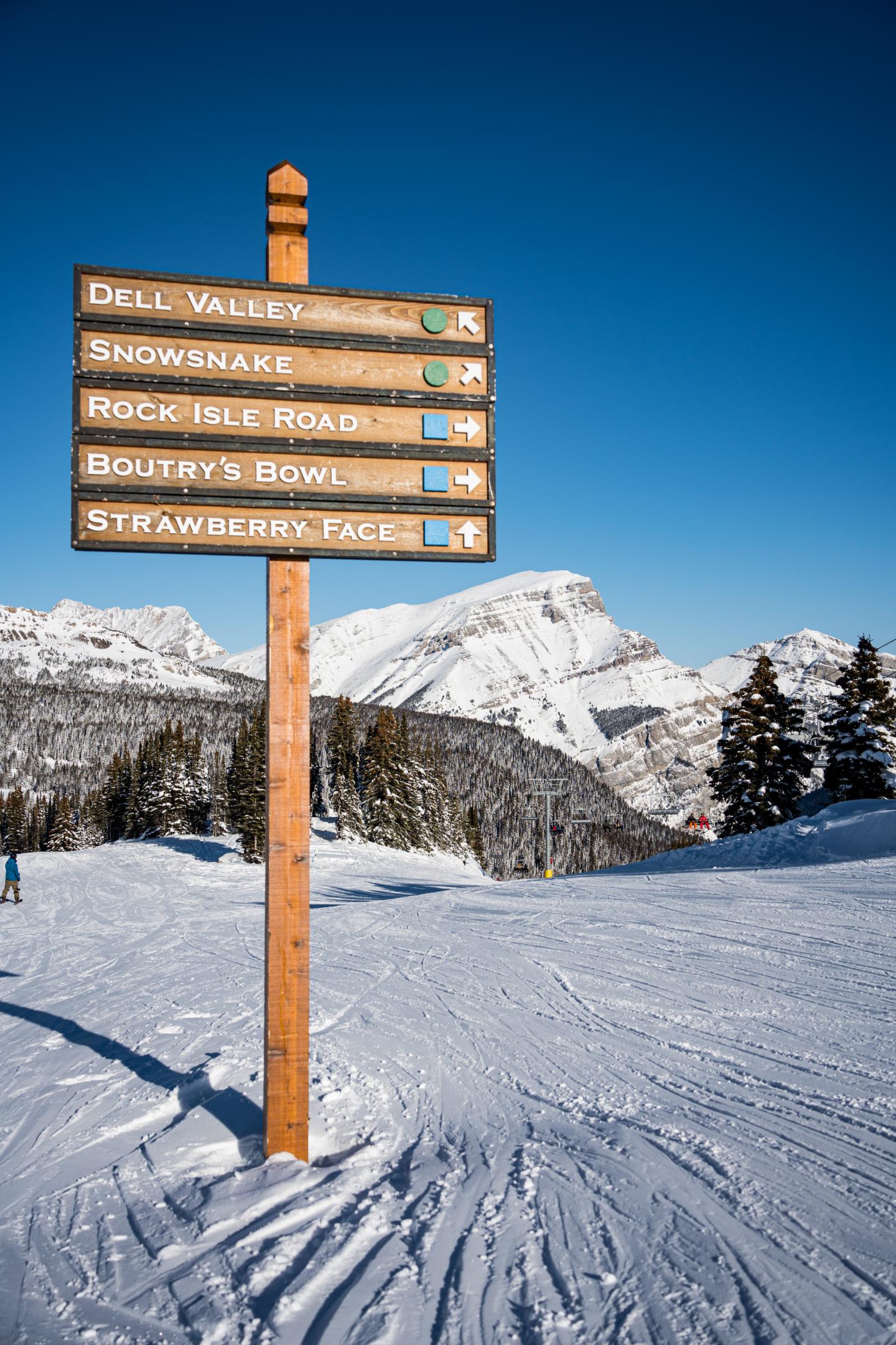 Ski trail signpost against snowy mountain backdrop under clear blue sky.