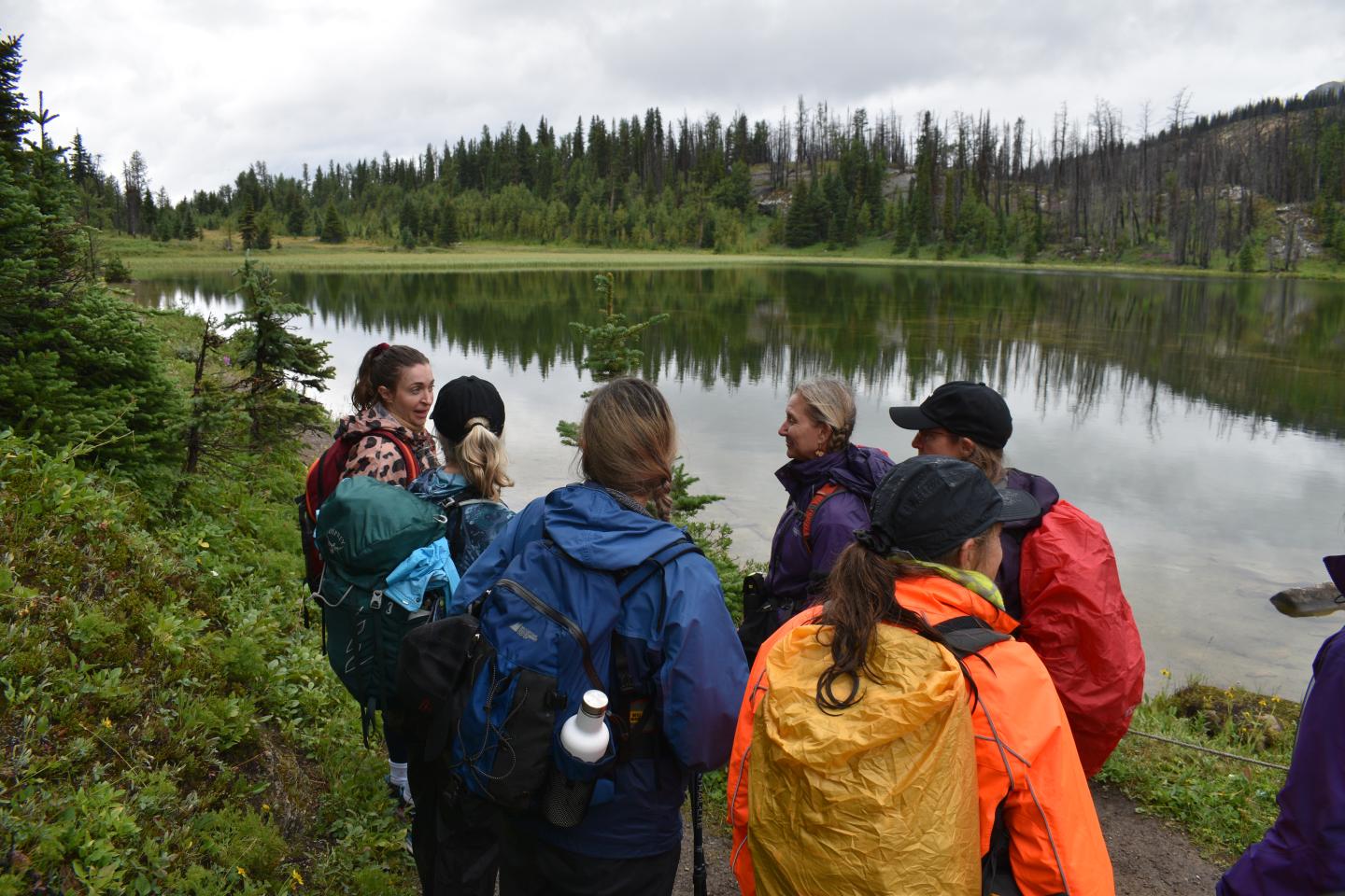Hikers gather by a lakeside under cloudy skies, surrounded by forest.