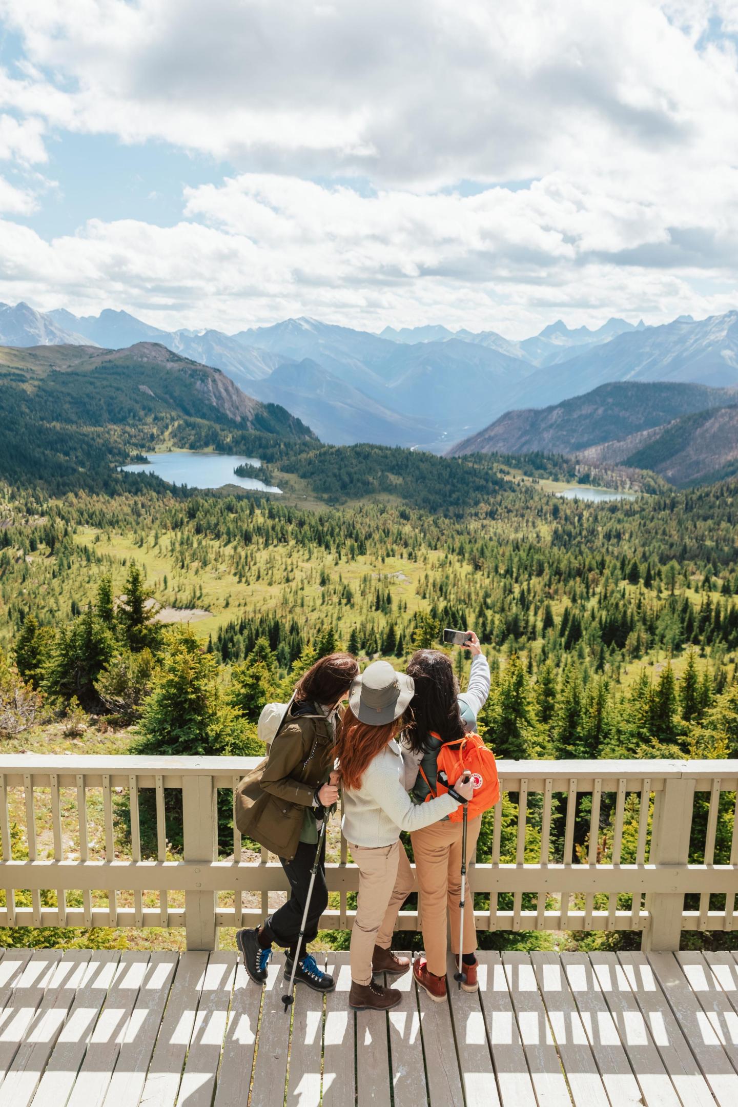 Friends taking a selfie on a scenic mountain viewpoint with lakes and forests.