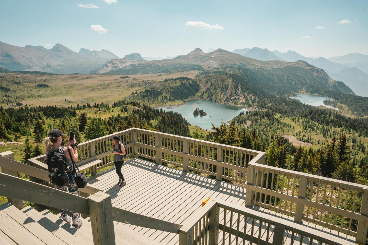 Wooden deck overlooking mountains, lakes, and forest under a clear sky. Two people taking photos.