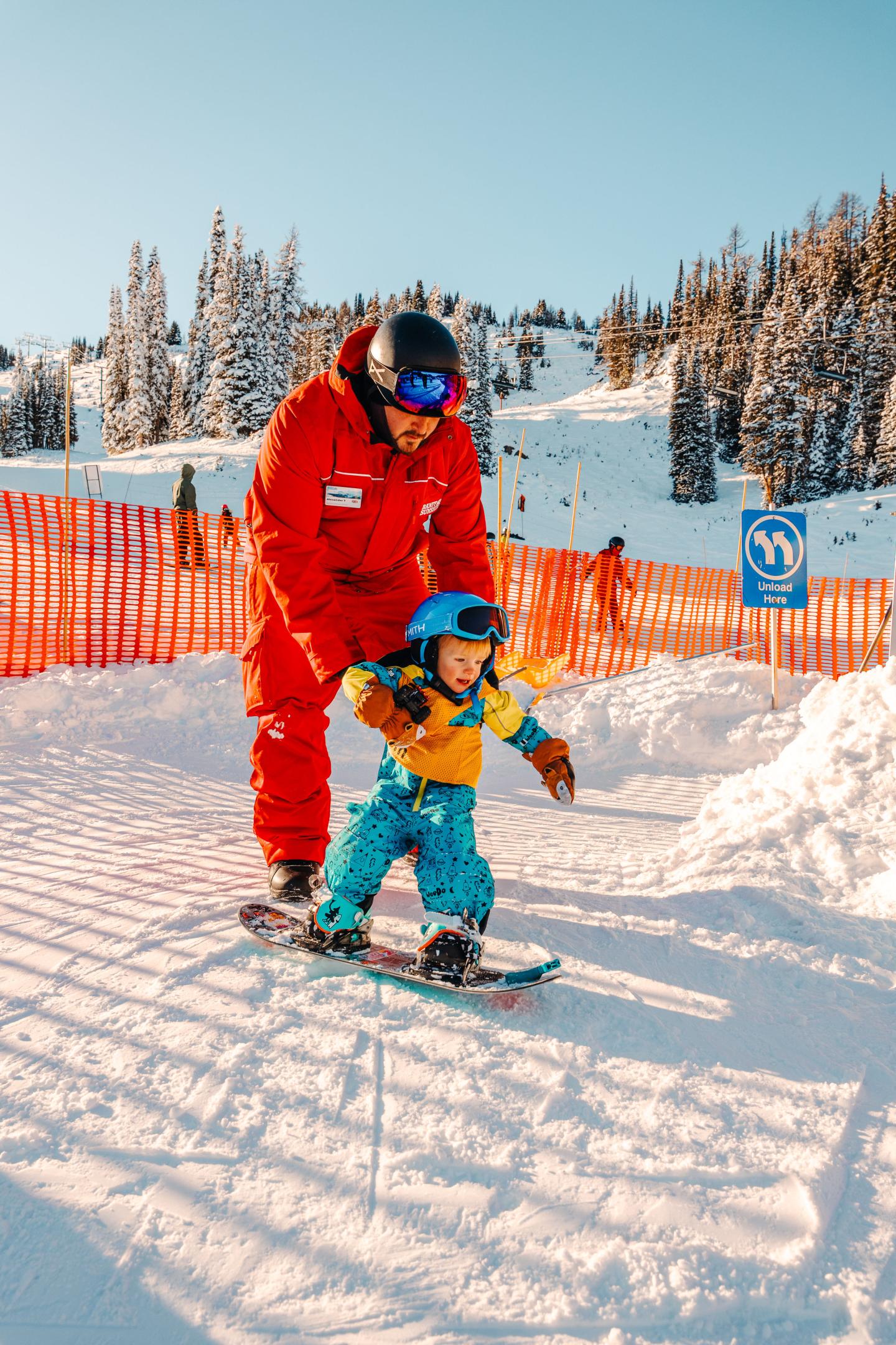 Instructor helps child snowboard on snowy slope, sunny day.