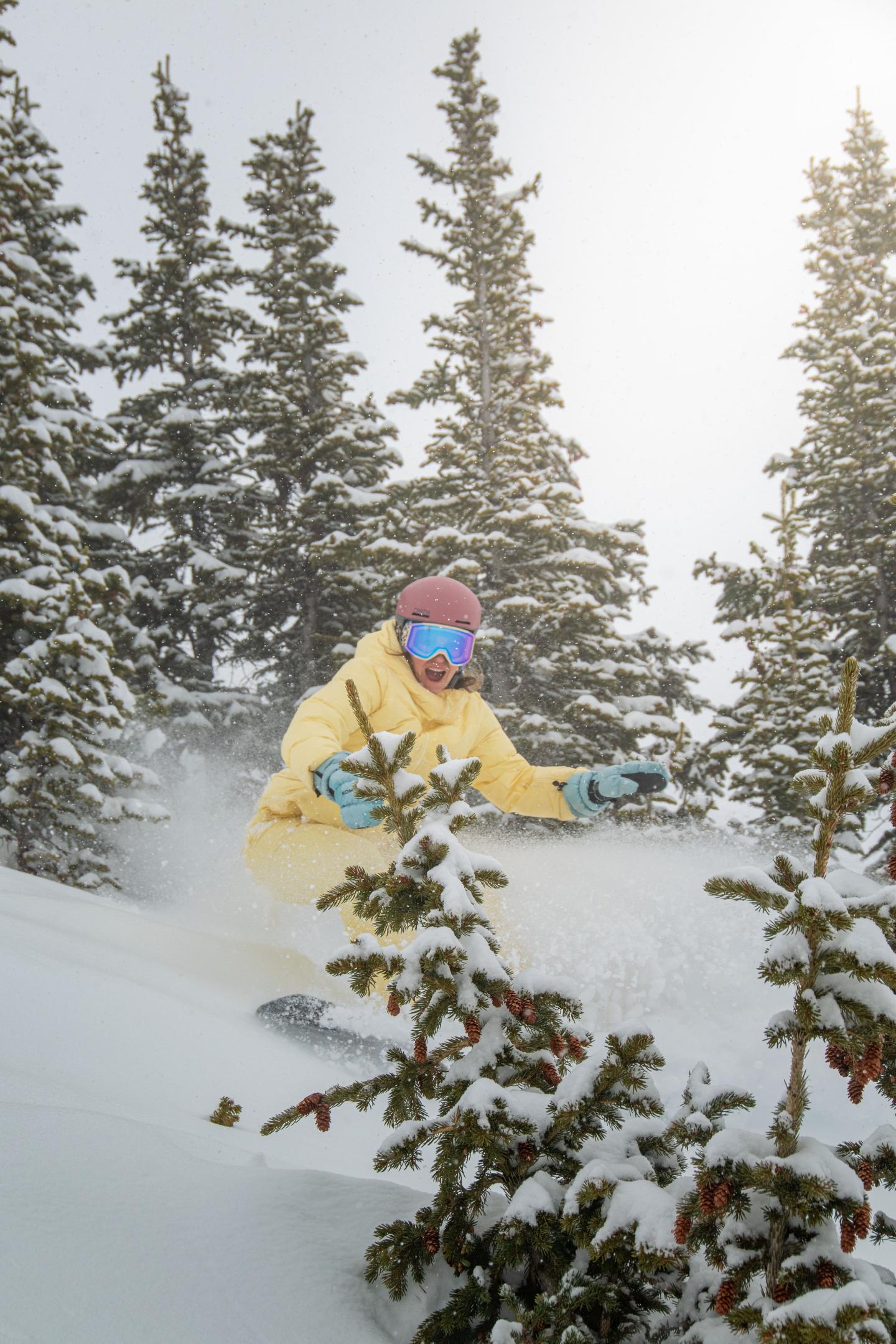 Snowboarder in yellow gear riding through snowy trees.