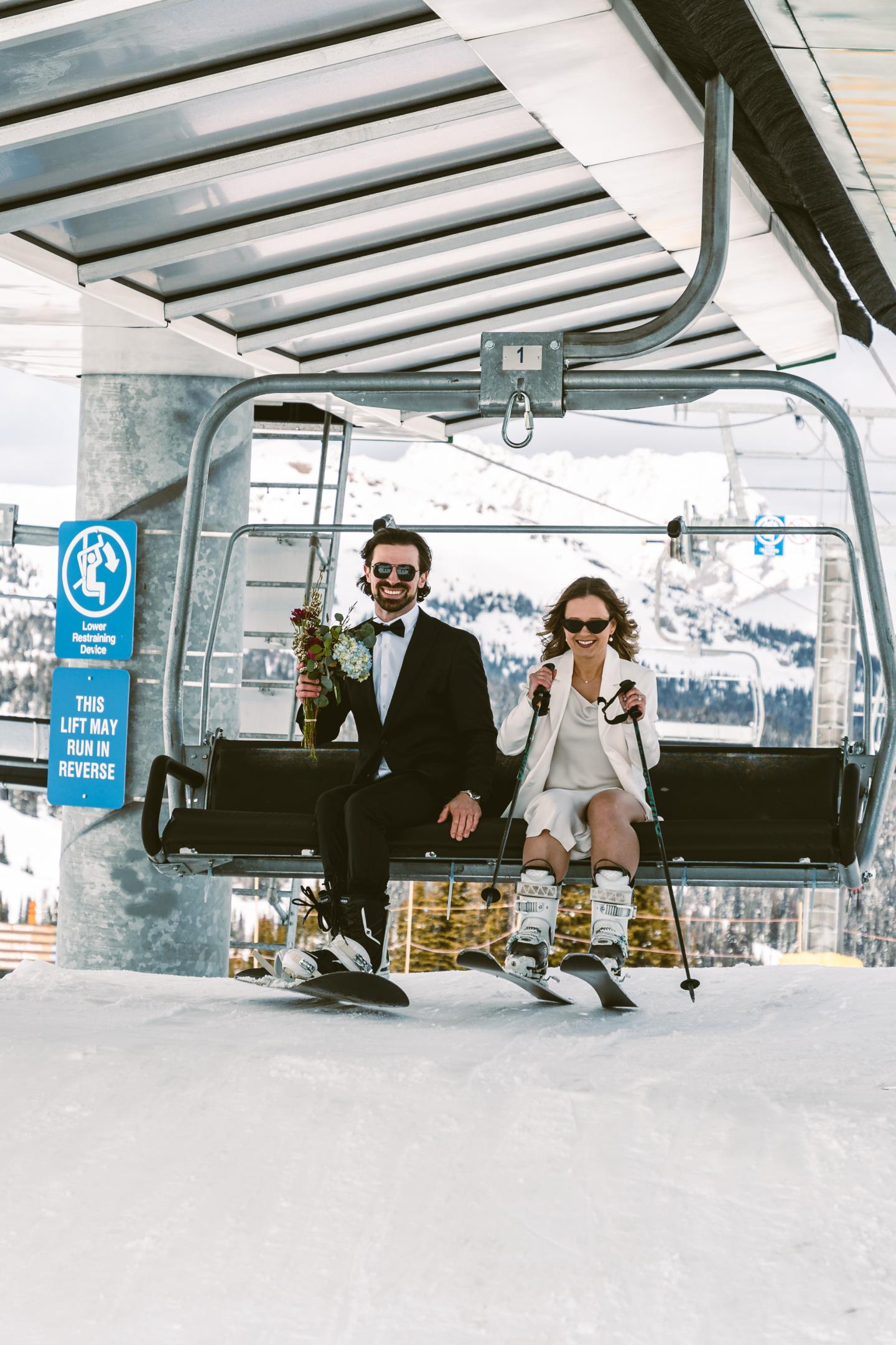 Couple in wedding attire on a ski lift, snowy mountain backdrop.