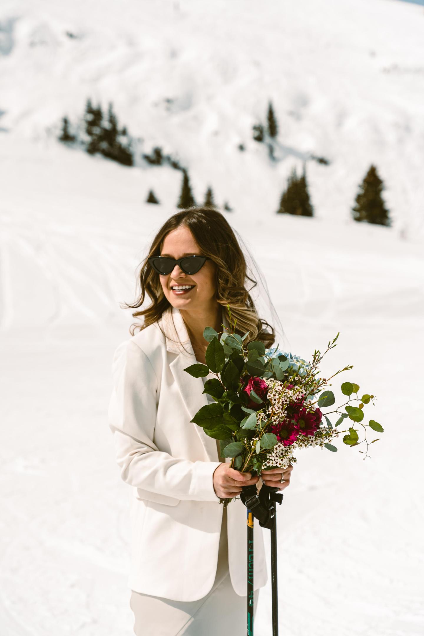 Bride in sunglasses holds bouquet, standing on snowy mountain.