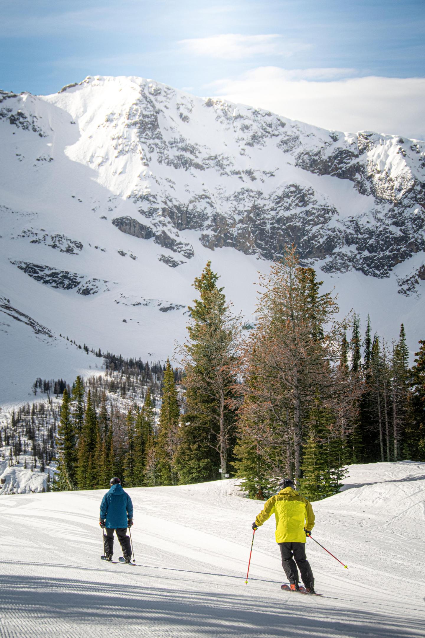 Two skiers glide down a snowy slope with mountains in the background.