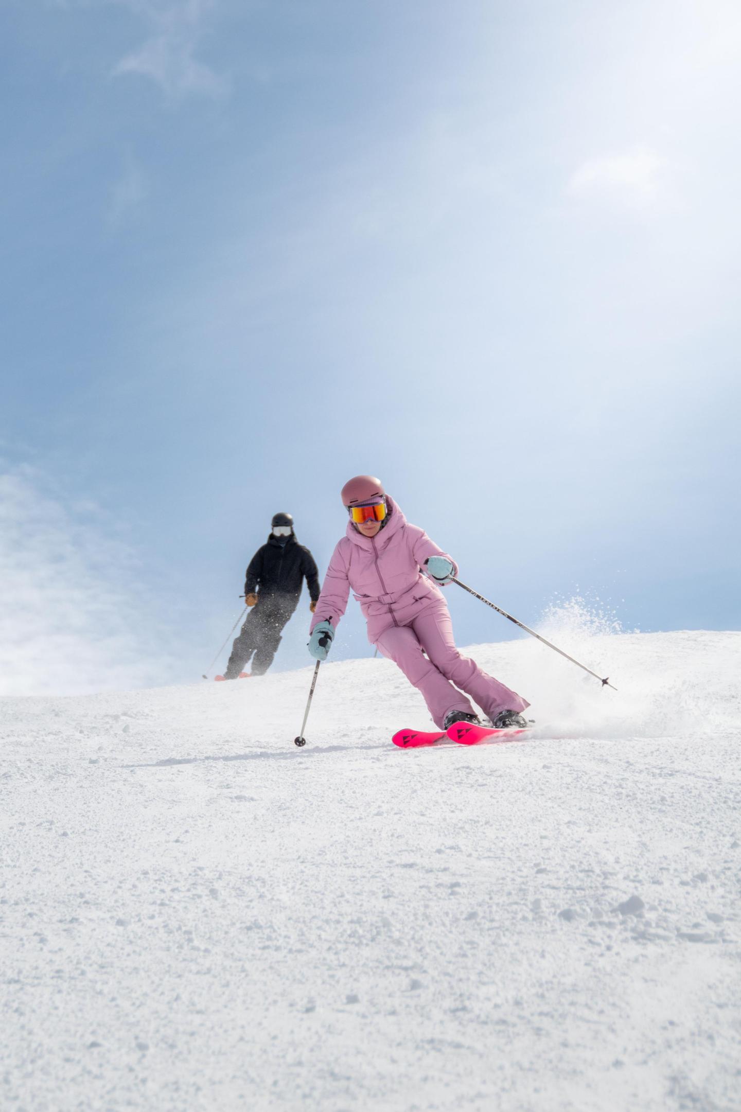 Two skiers in colorful gear skiing down a snowy slope under a clear sky.