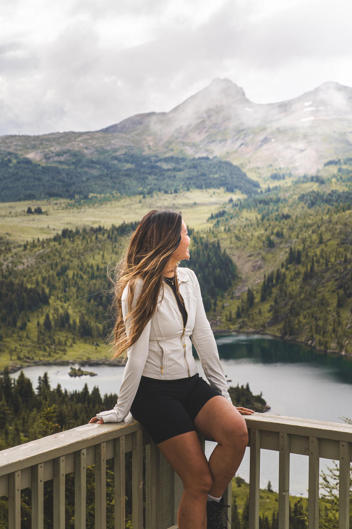 Woman sitting on a railing, gazing at mountains and lake scenery.