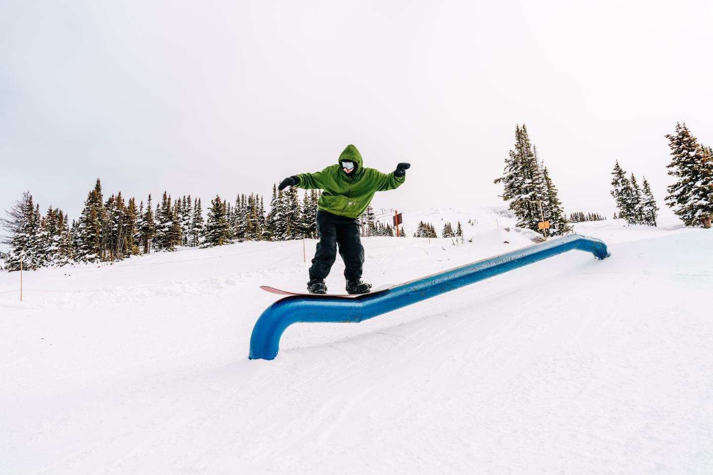 Snowboarder in a green jacket sliding on a blue rail in a snowy landscape.