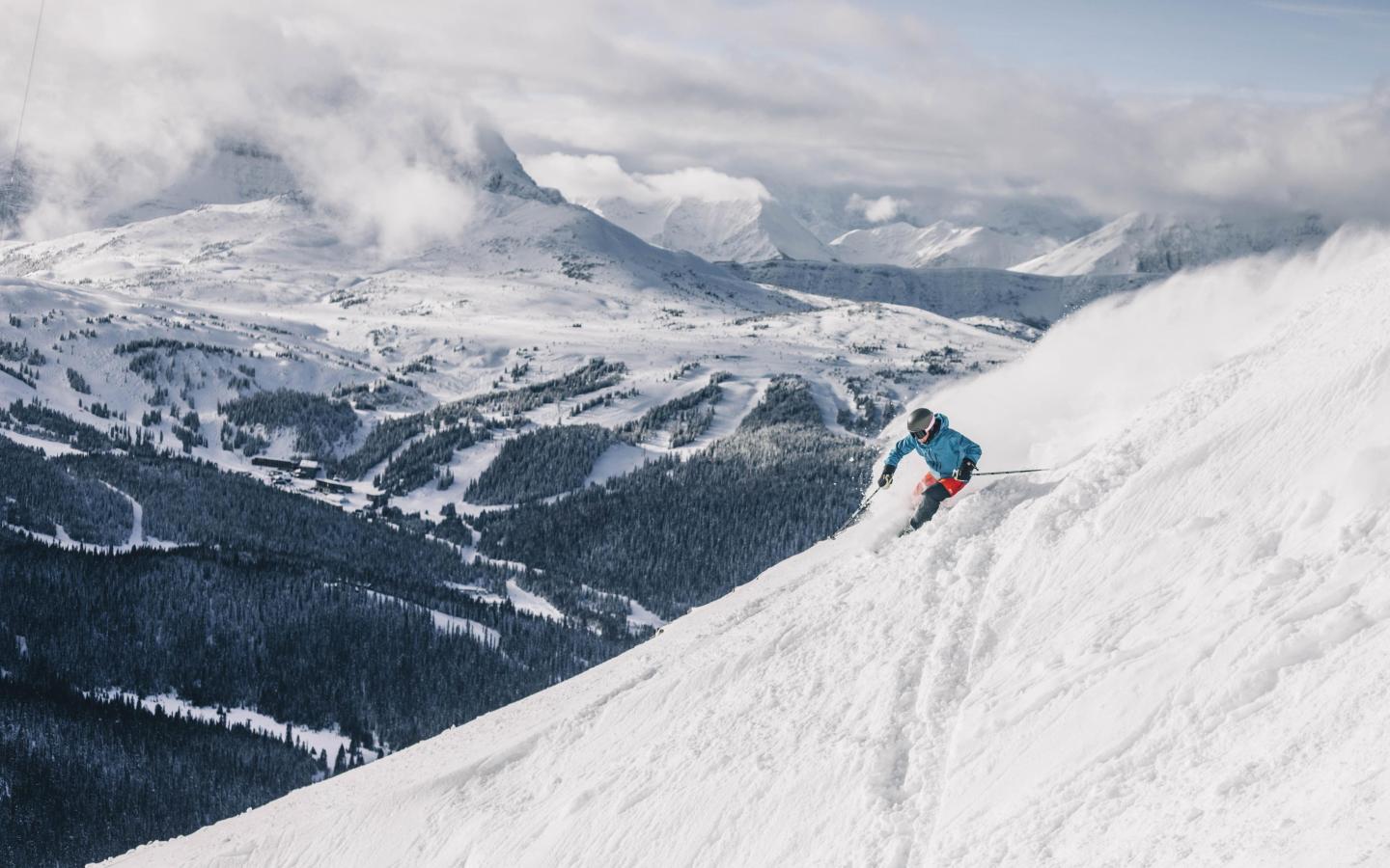 Skier in blue jacket descends snowy mountain slope with distant snowy peaks.