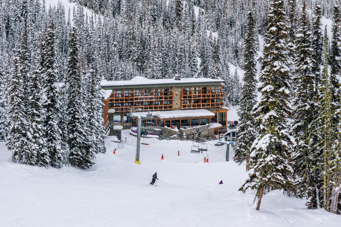 Snowy landscape with skiers and a lodge among tall pine trees.