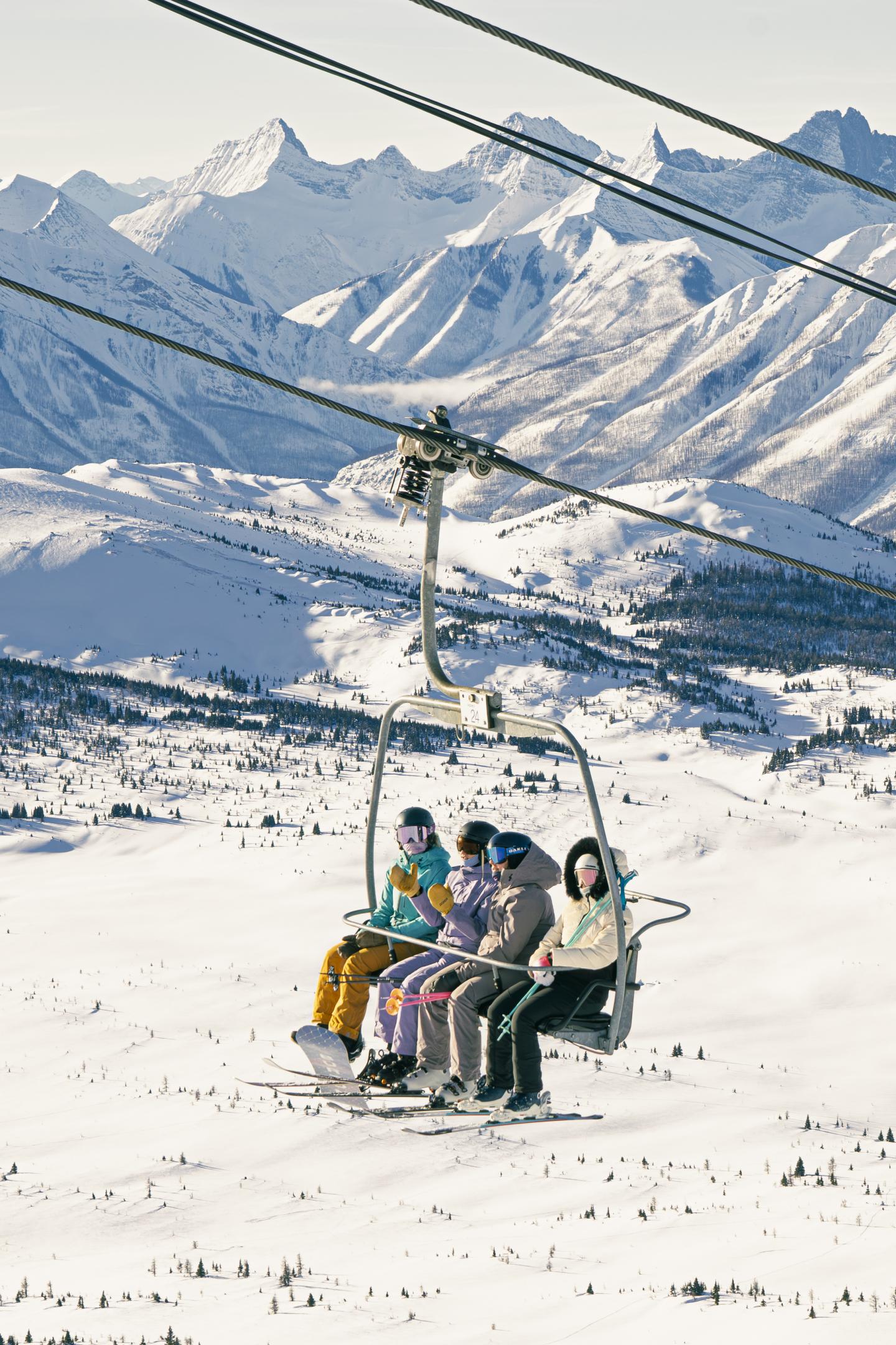 Ski lift with four people, snow-covered mountains in the background.