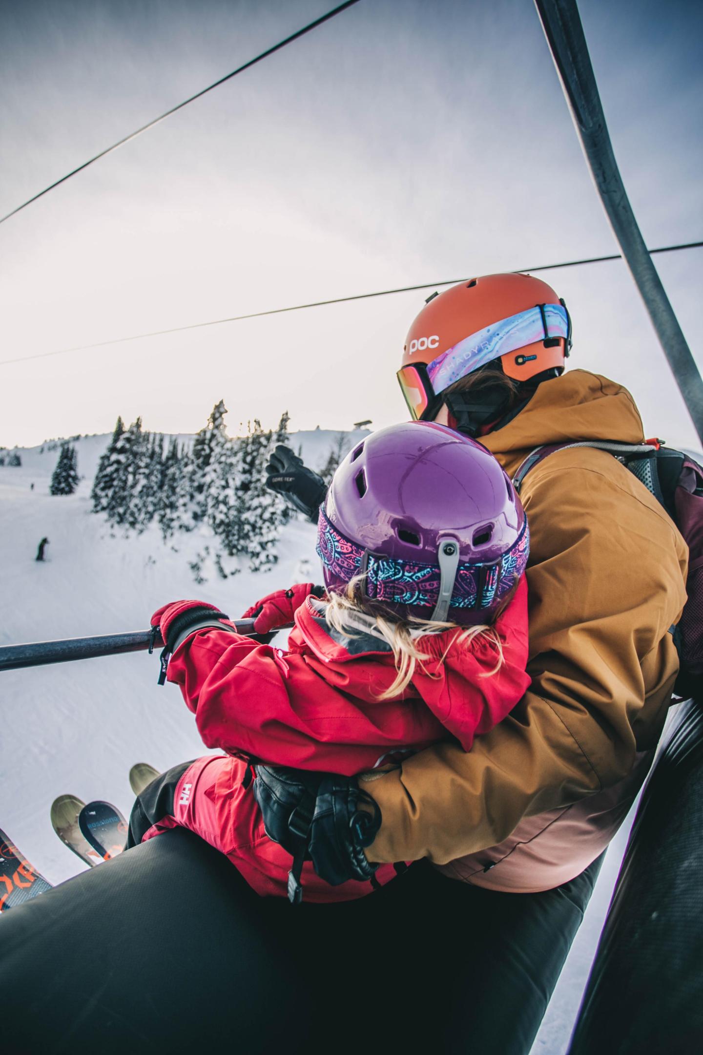 Parent and child on a ski lift, wearing helmets and winter clothing.