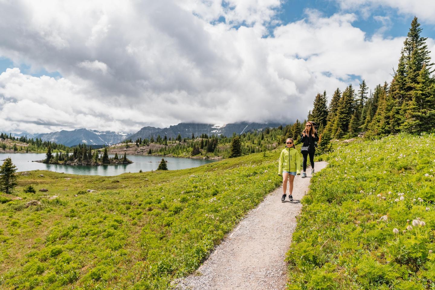 Hikers walking on a scenic trail near a lake, surrounded by mountains and trees.