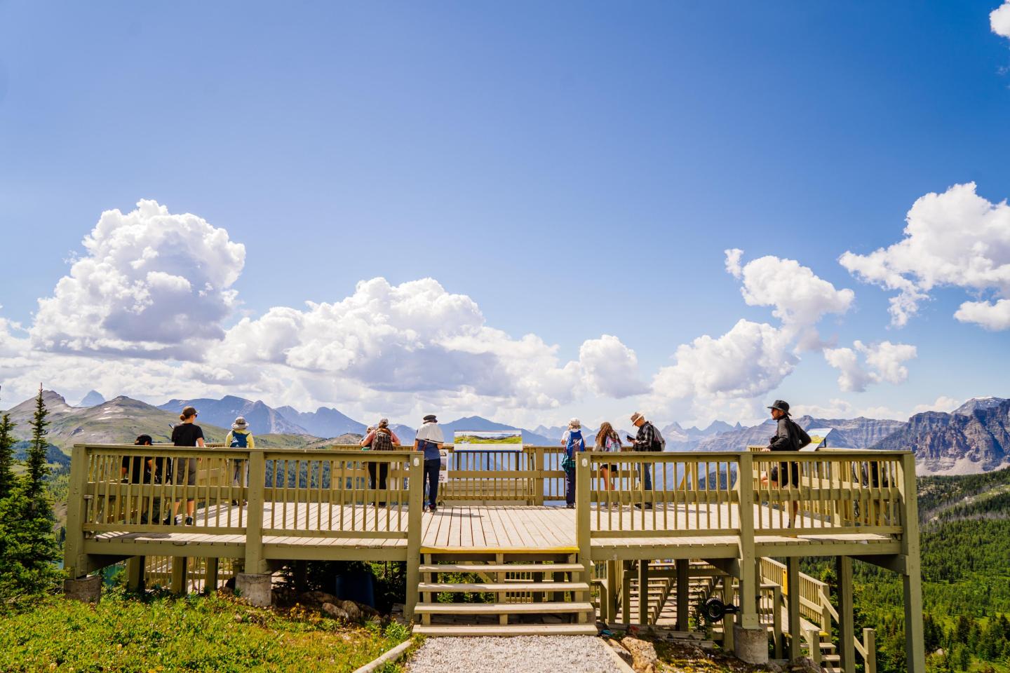 Wooden lookout deck with people, surrounded by mountains and a bright blue sky with fluffy clouds.
