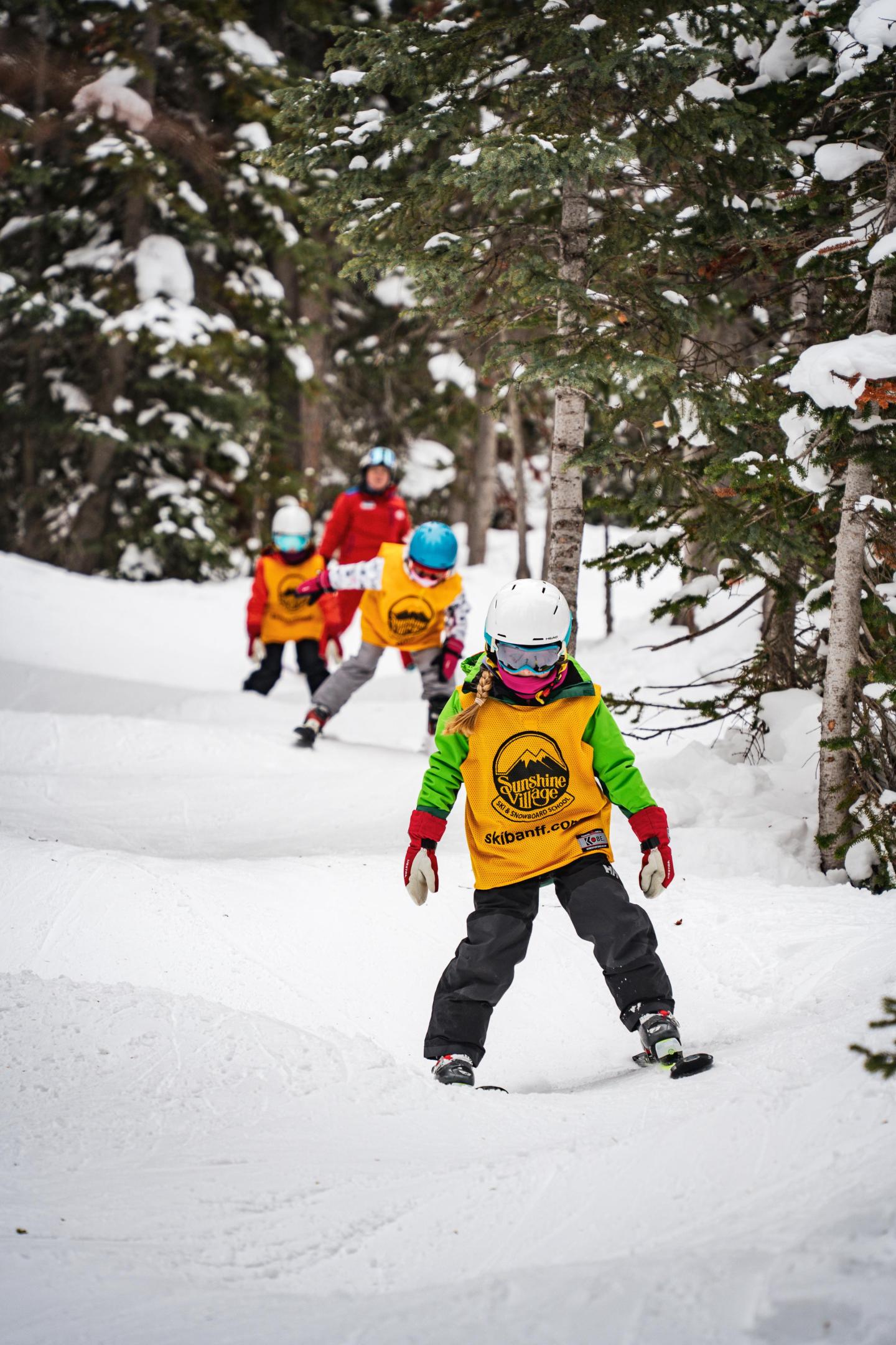 Children snowboarding down a snowy hill, wearing helmets and yellow vests.