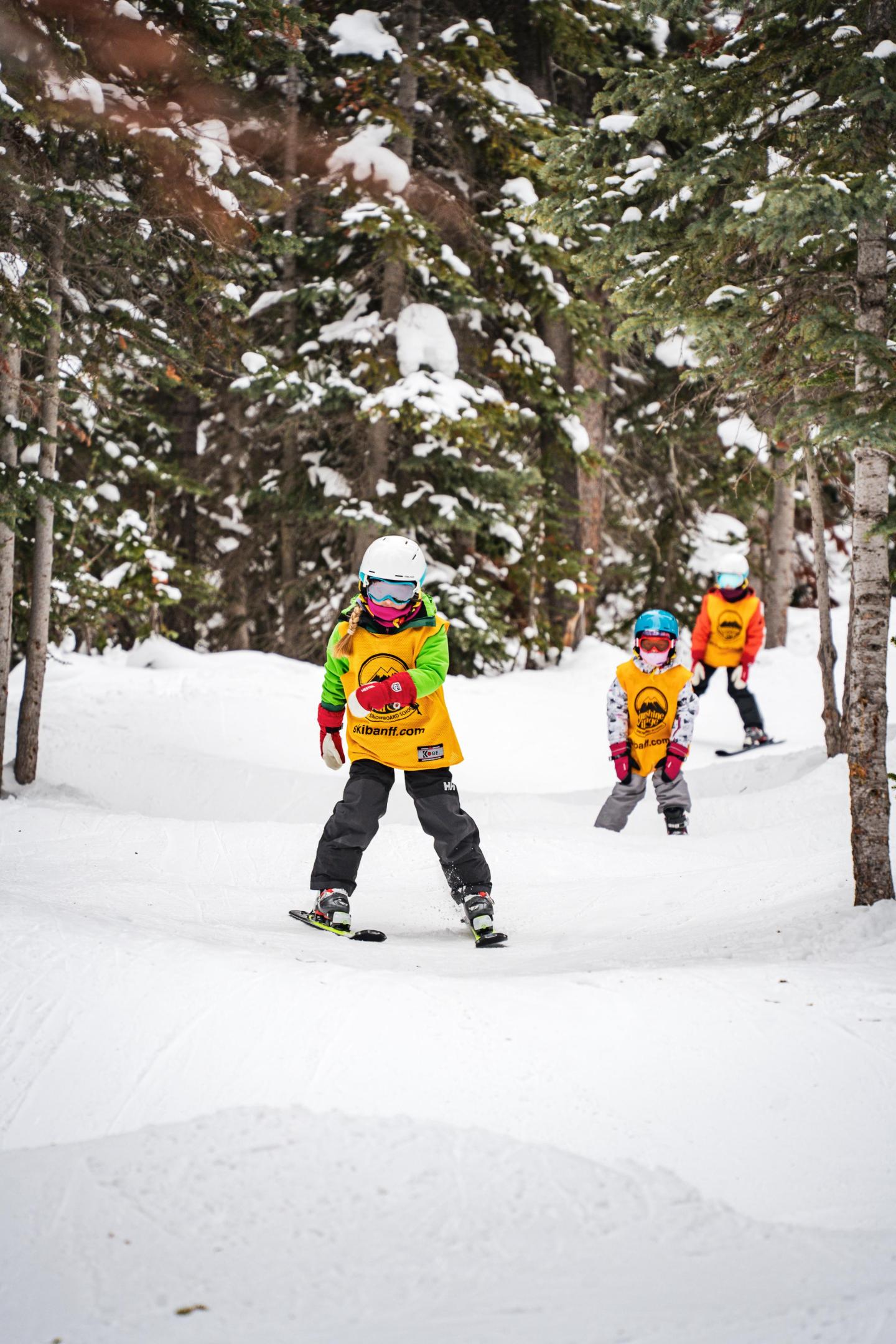 Children skiing through a snowy forest, wearing yellow vests and helmets.