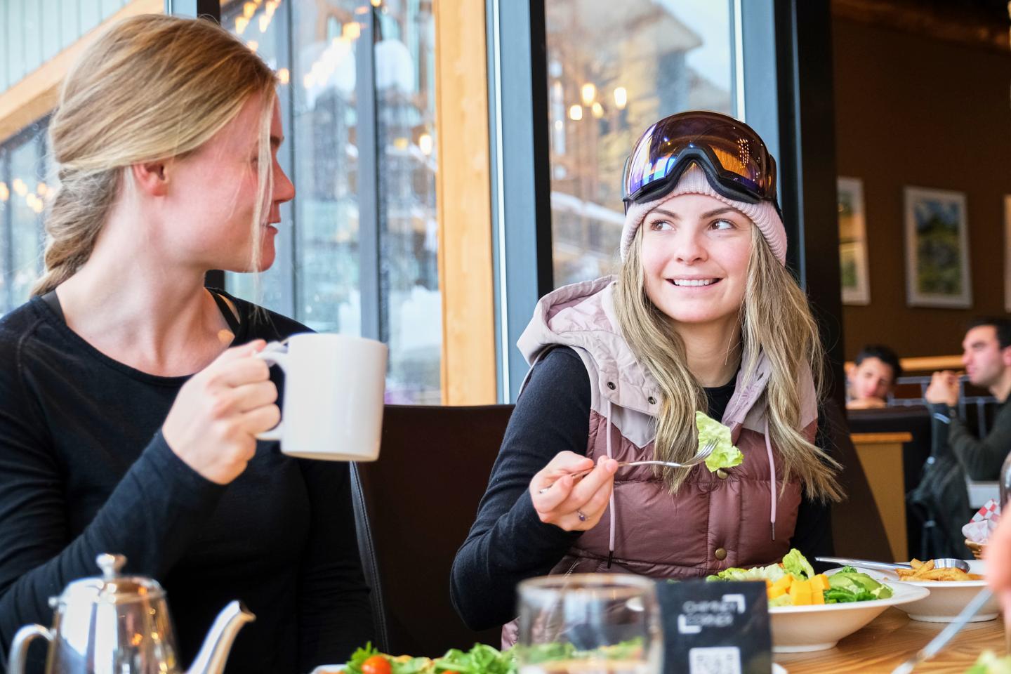 Two women enjoying a meal in a cozy cafe, one wearing ski goggles.