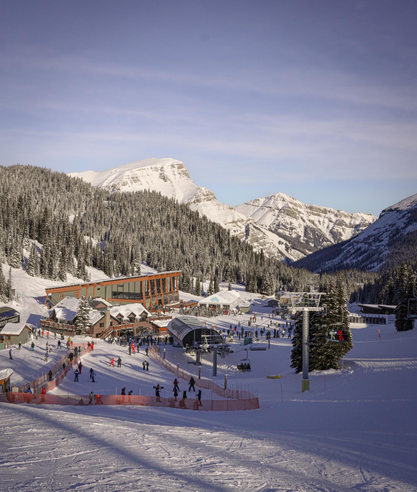 Snowy mountain ski resort with skiers, a lodge, and chairlift under a blue sky.