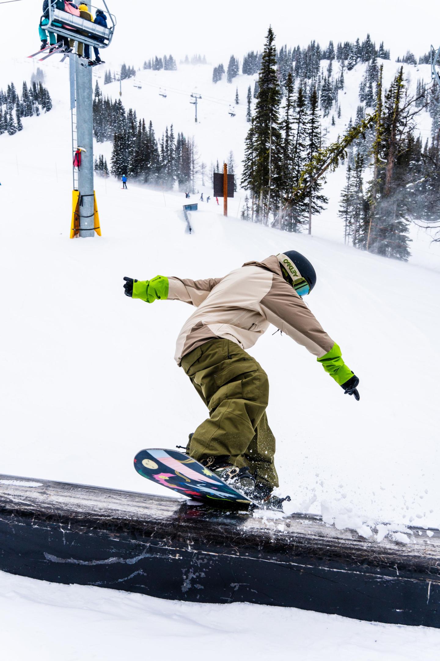 Snowboarder in beige jacket grinding on a rail in a snowy mountain setting.