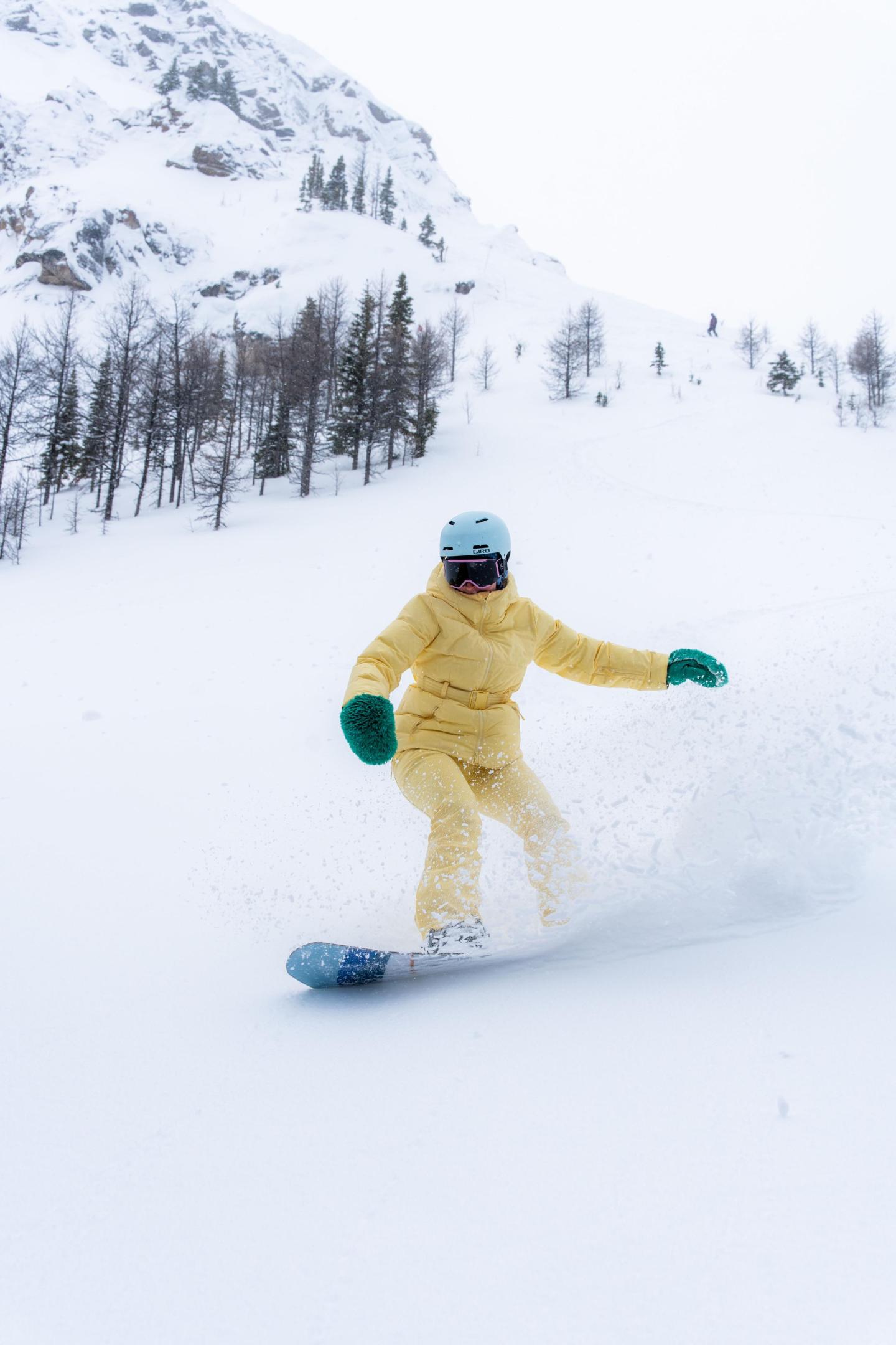 Snowboarder in yellow outfit glides down snowy mountain, trees in background.