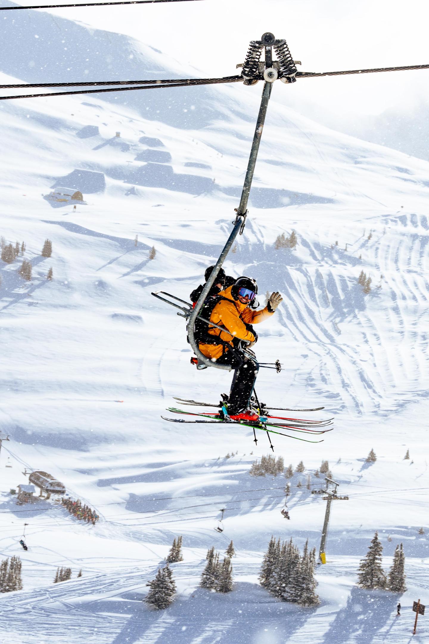 Skier in orange jacket on ski lift over snowy slopes.