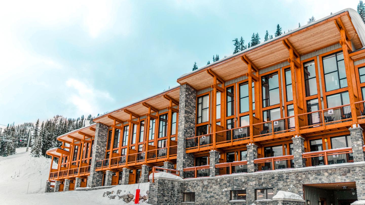 Modern building with stone and wood facade, snowy landscape, and pine trees.