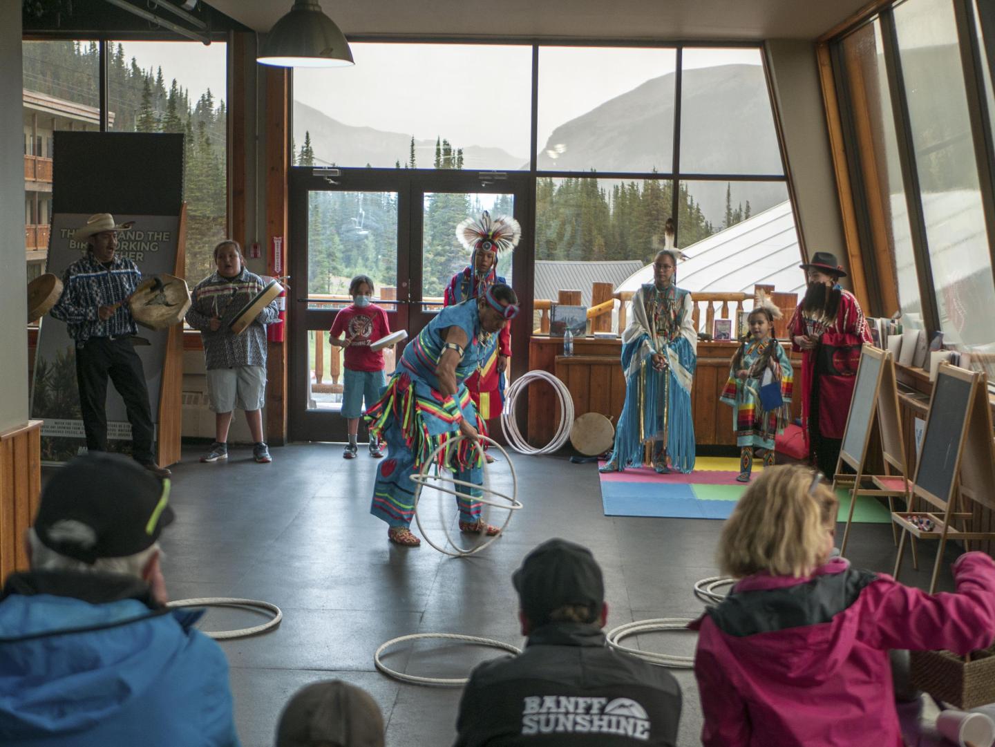 Dancers in colorful outfits perform with hoops indoors, audience watches.