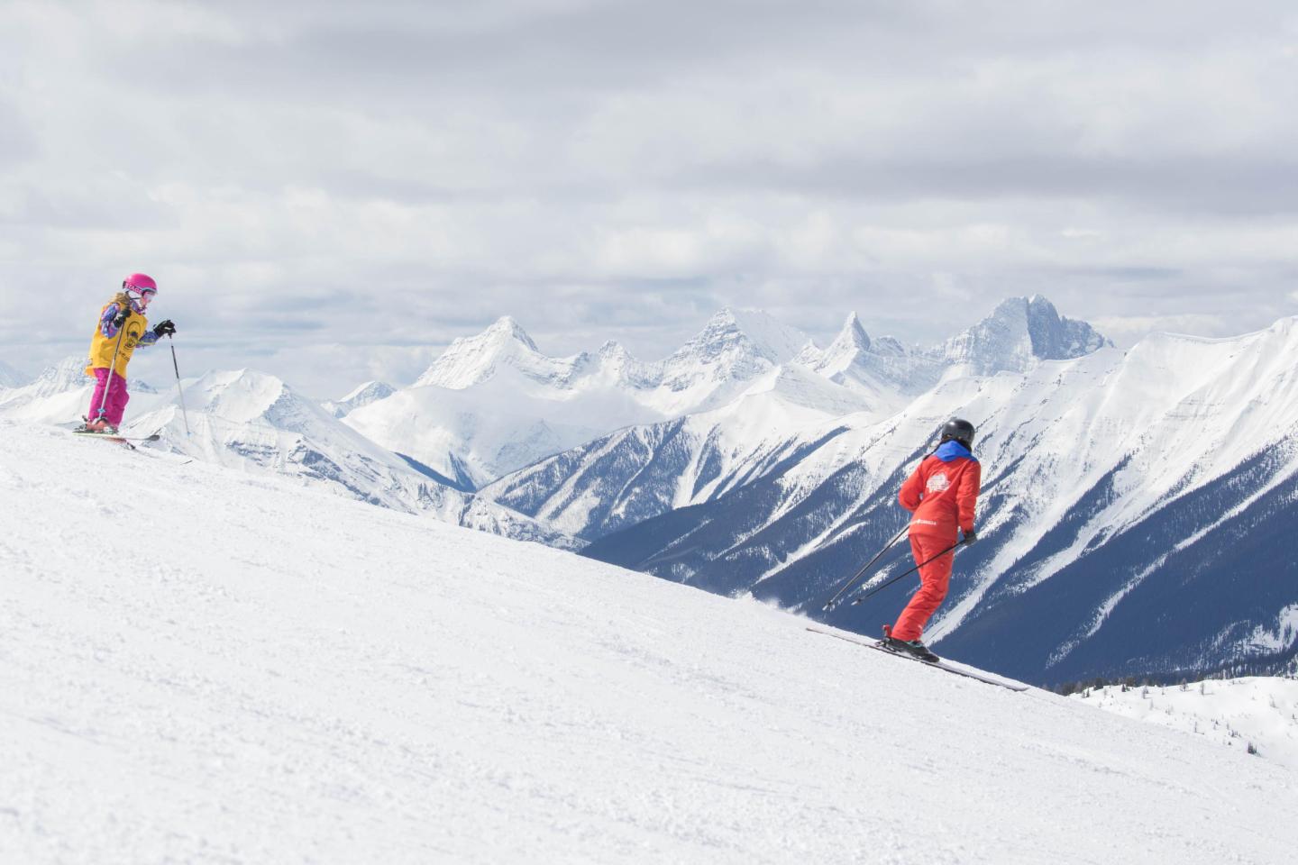 Two skiers in colorful outfits on a snowy mountain slope with cloudy, snowy peaks.