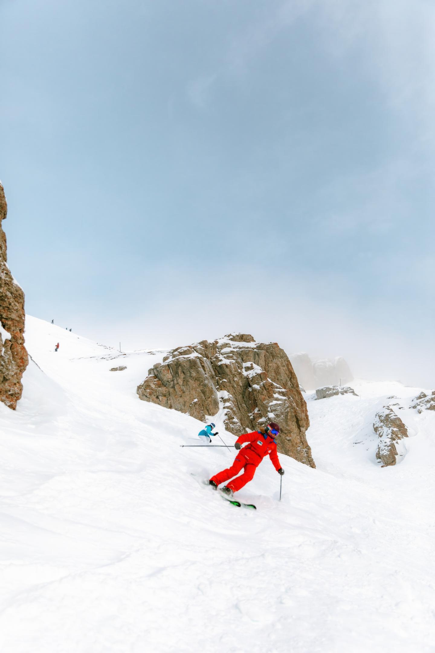 Skier in red suit descends snowy slope with rocky backdrop.