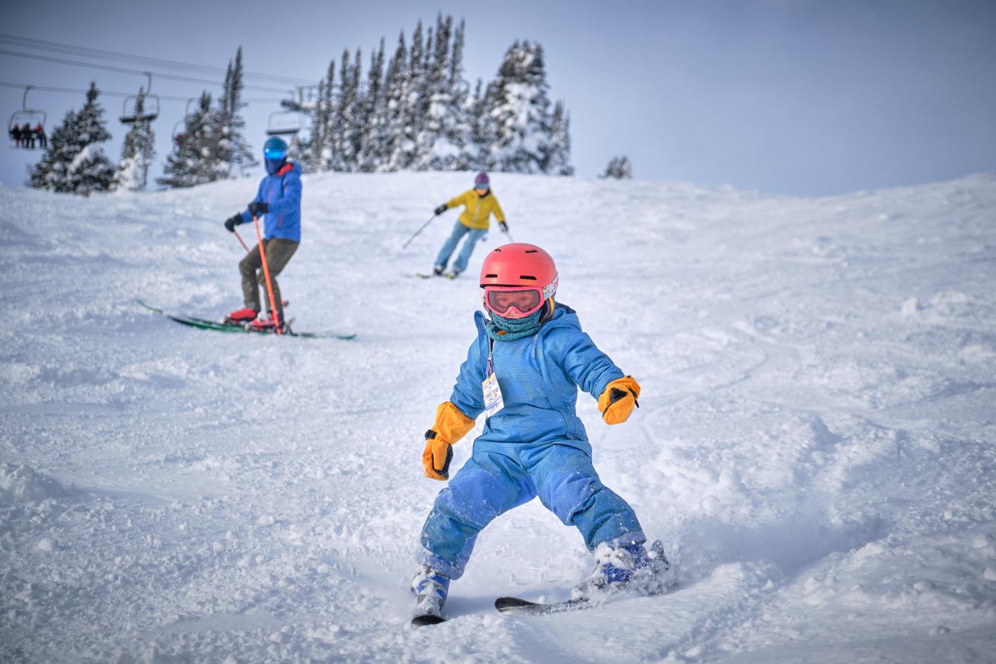 Child in blue snowsuit skiing downhill, with another skier behind.