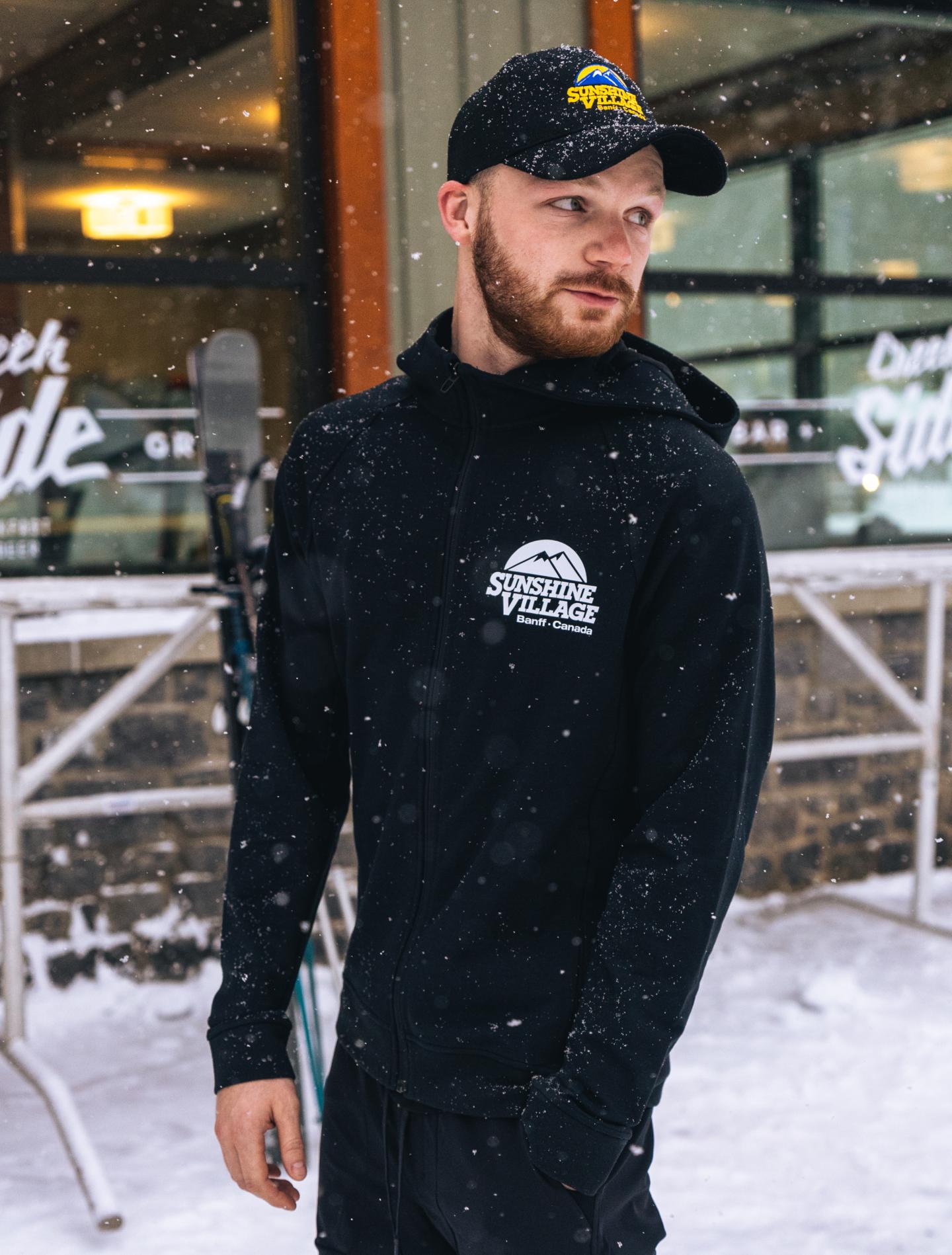 Man in a winter jacket and cap standing in falling snow outside a café.