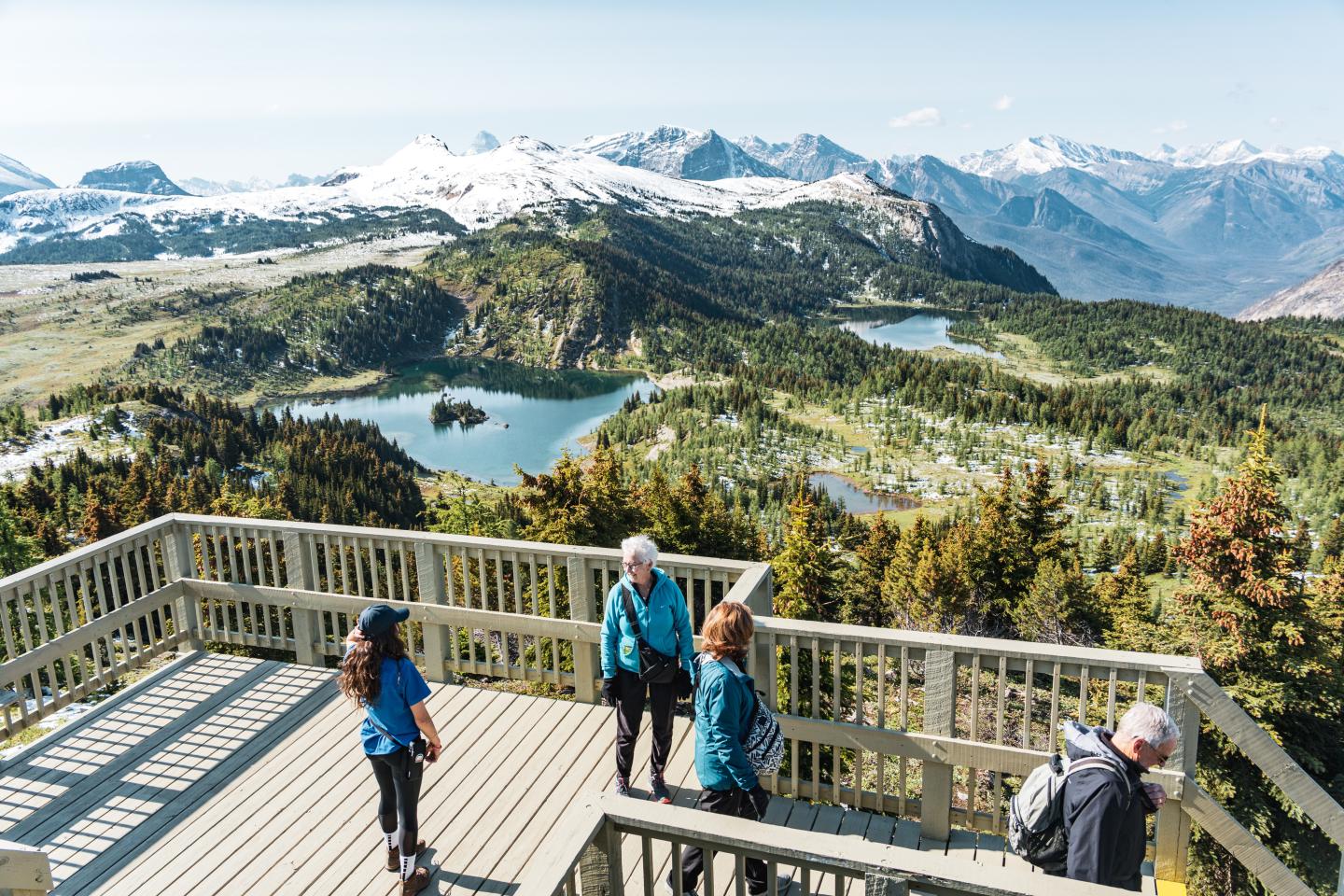 People standing on a wooden platform overlooking mountains and lakes under a clear sky.