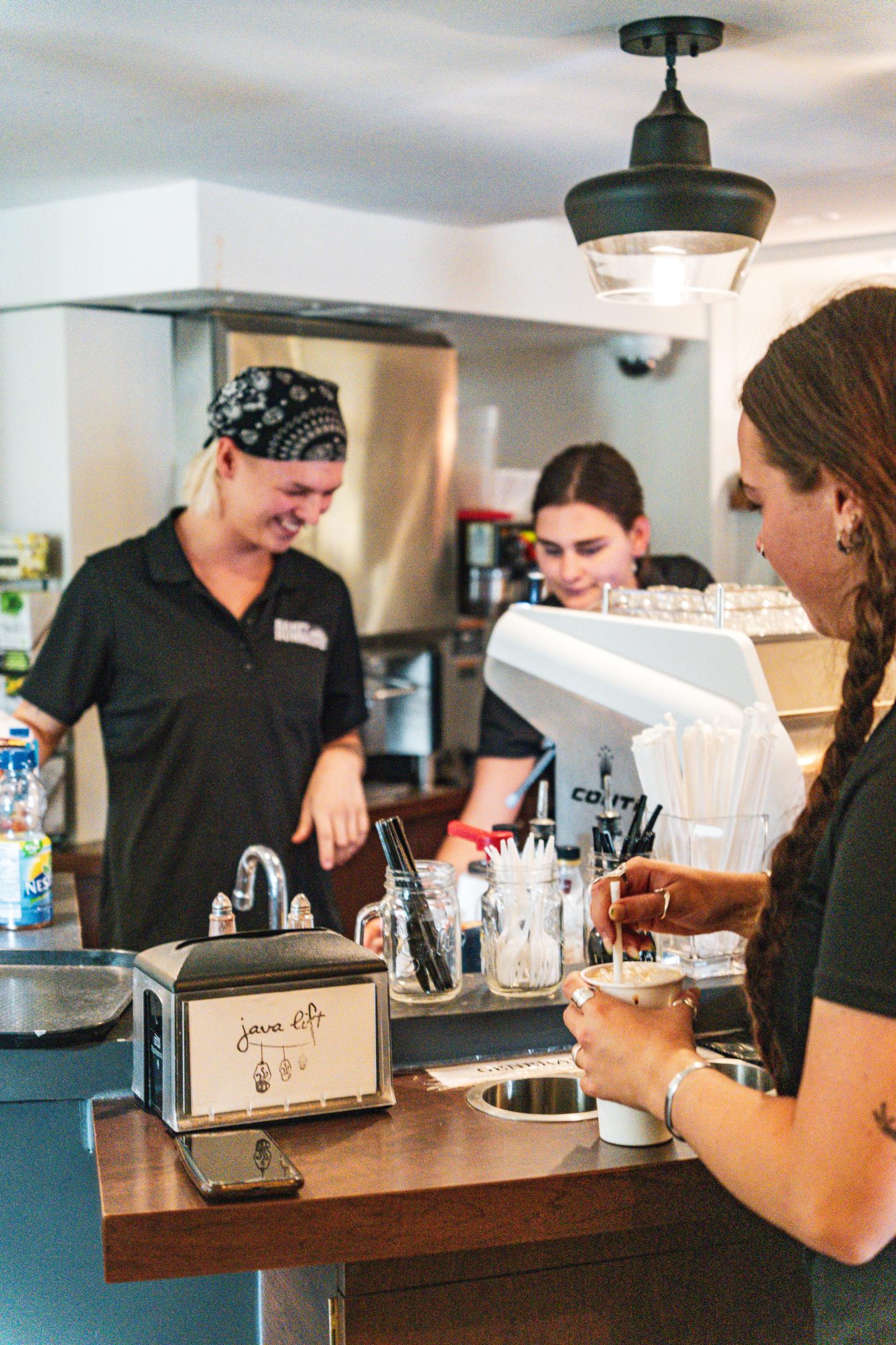 Three baristas working behind a counter, smiling and preparing drinks.