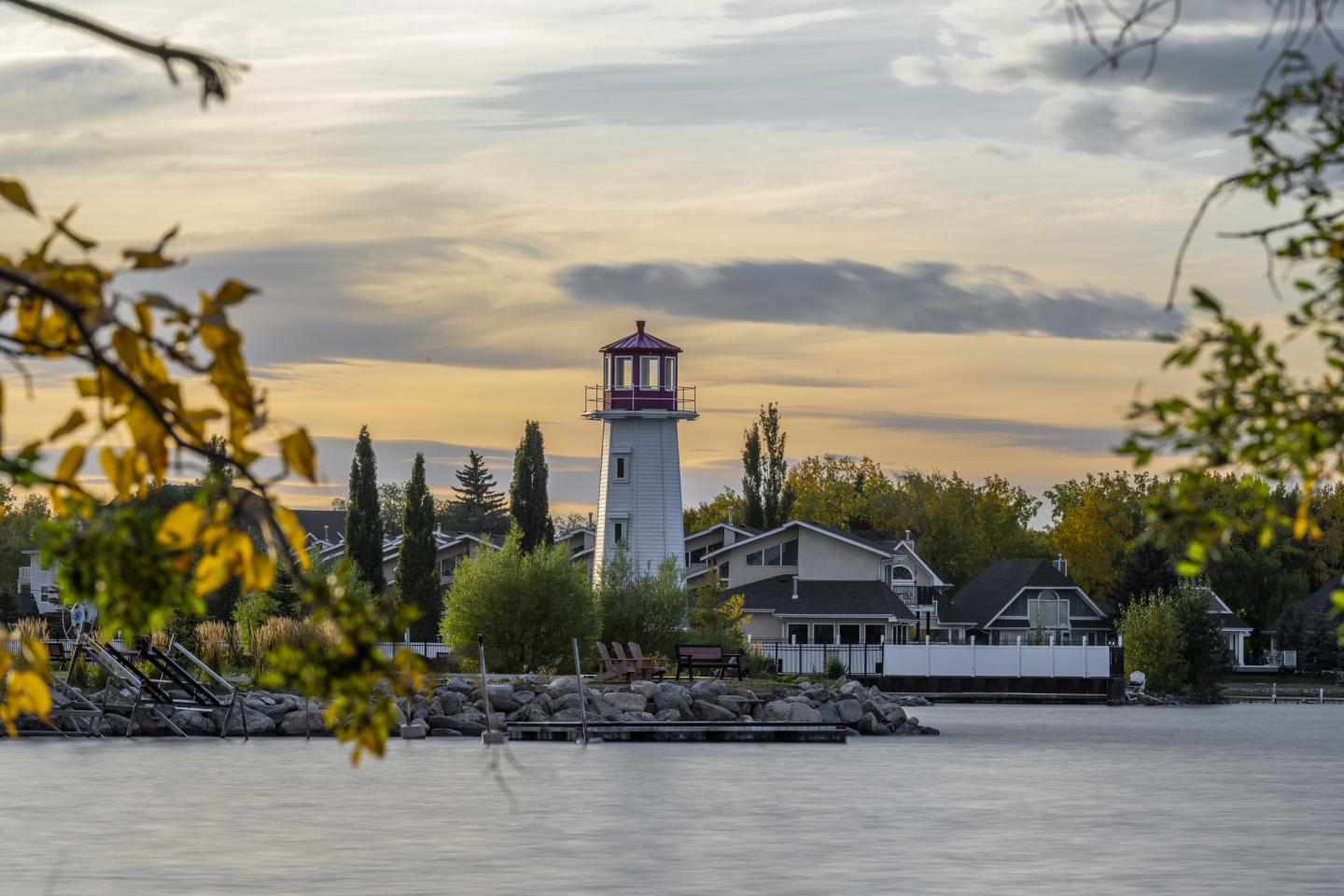 Lakeside lighthouse and houses at sunset, framed by autumn leaves.