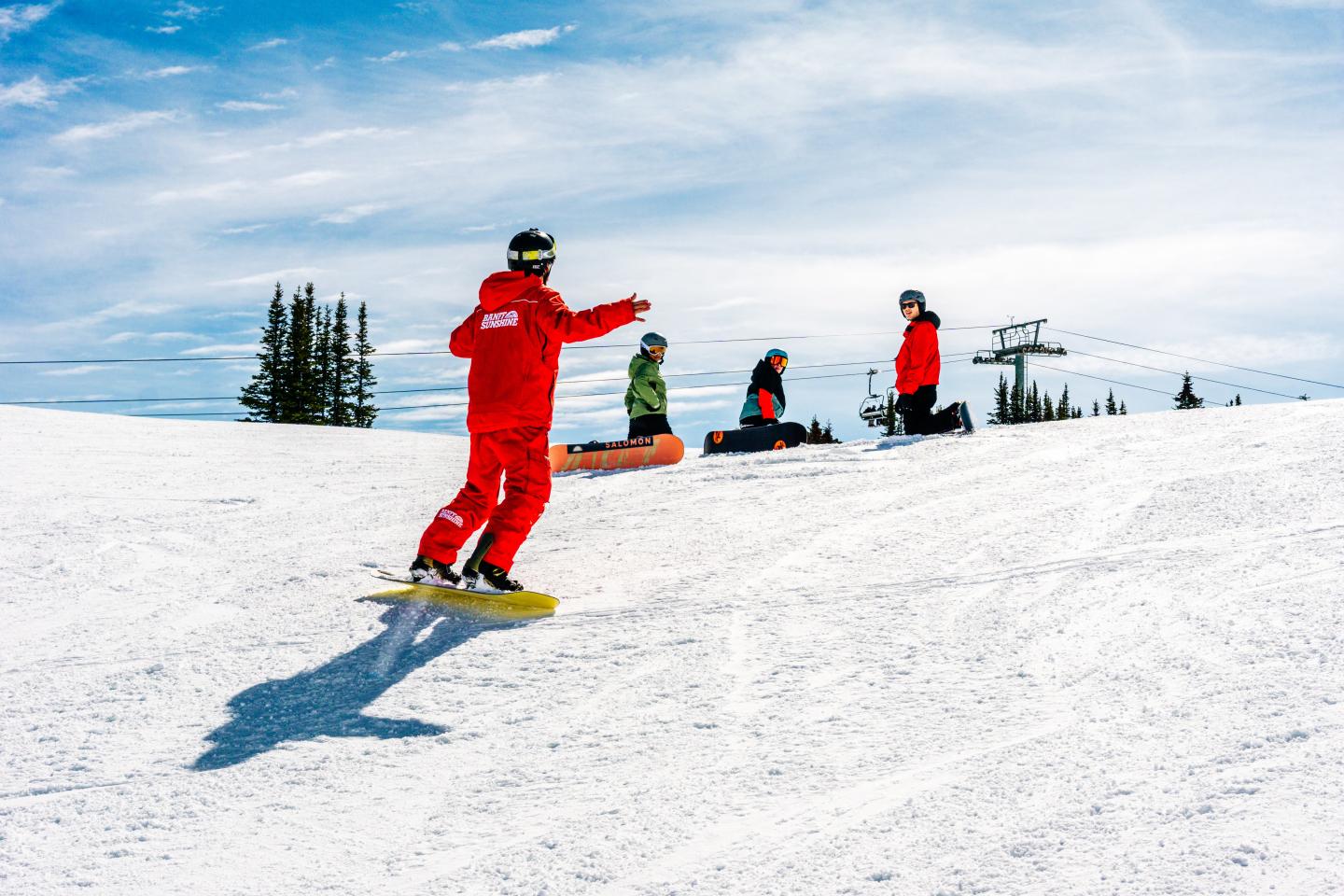 Snowboarders in red gear on a snowy hill under a clear blue sky.