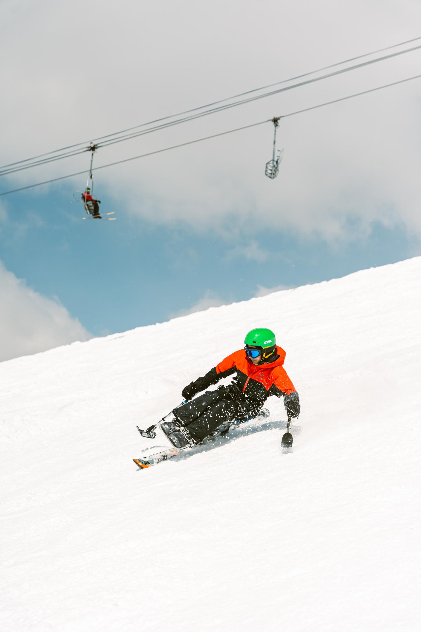 Sit skier in orange jacket descends snowy slope under cloudy sky.
