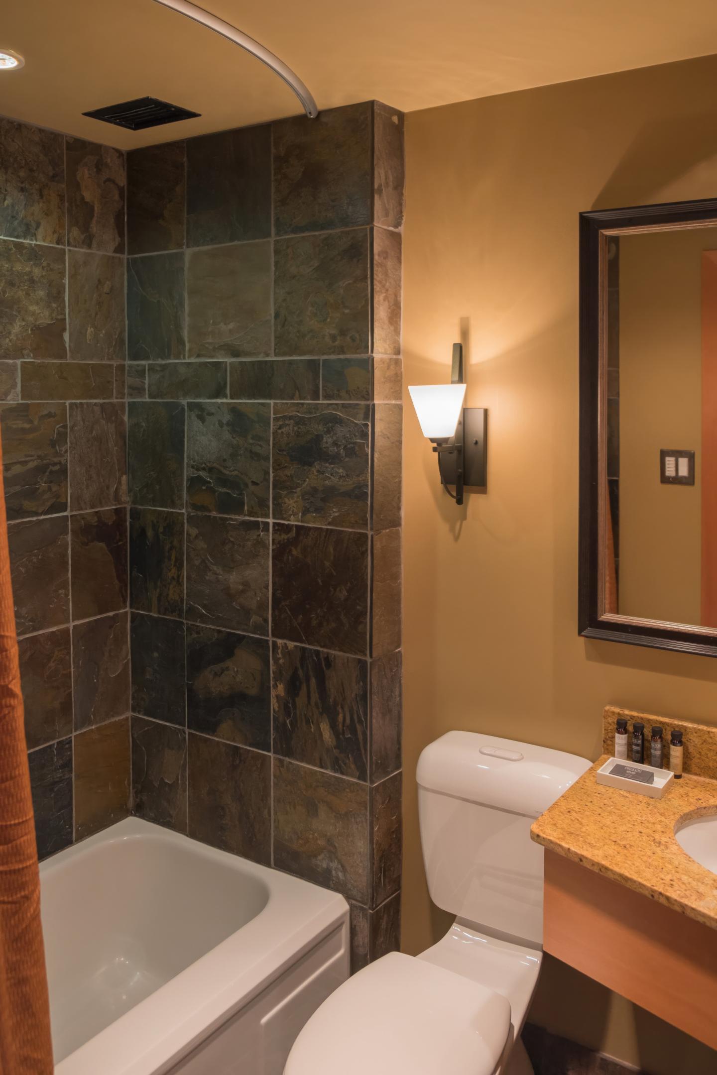 Warm bathroom with tiled shower, white tub, vanity, and wall sconce.