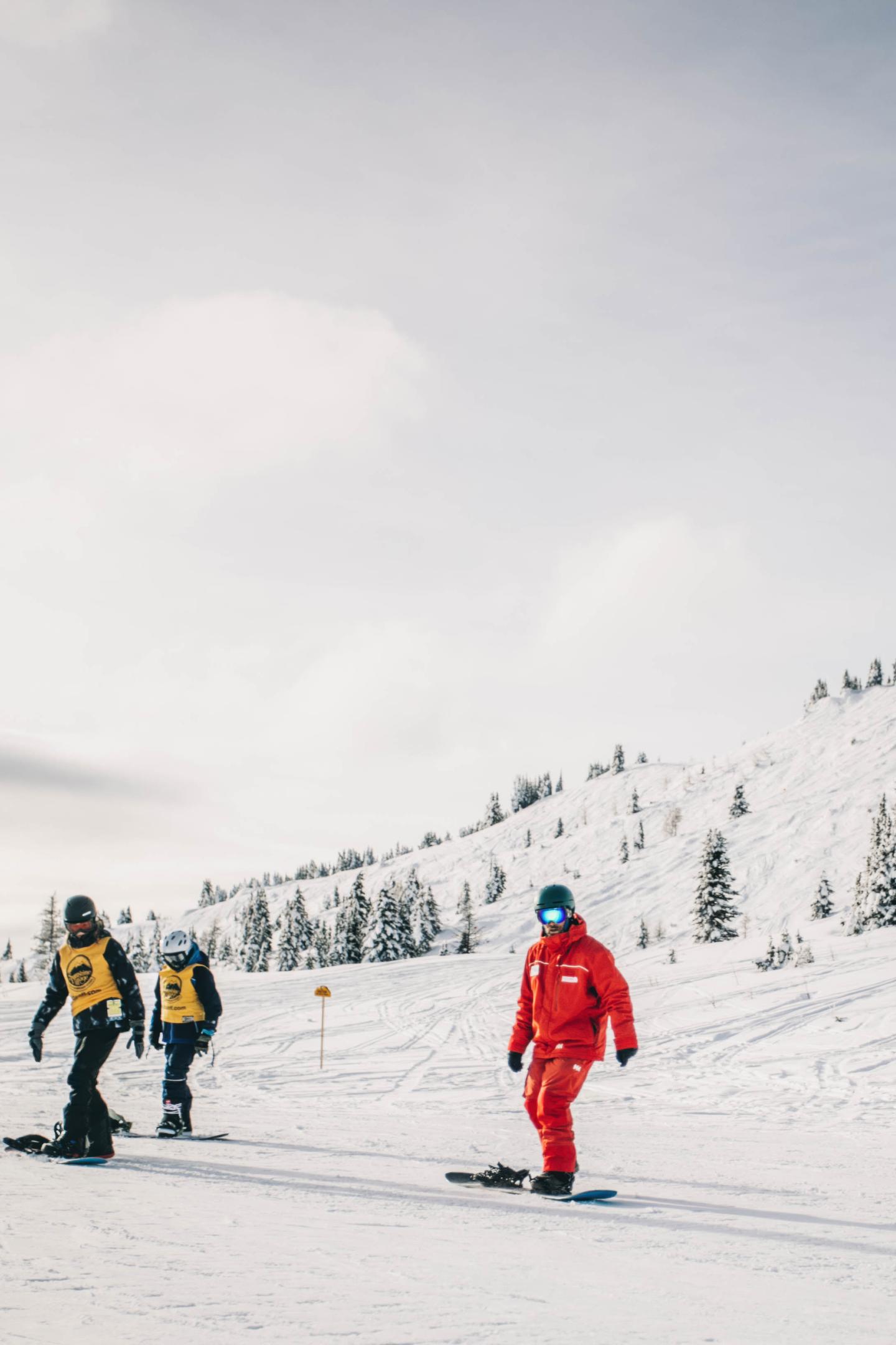 Snowboarders in snowy landscape with pine trees, one wearing red, others in yellow vests.
