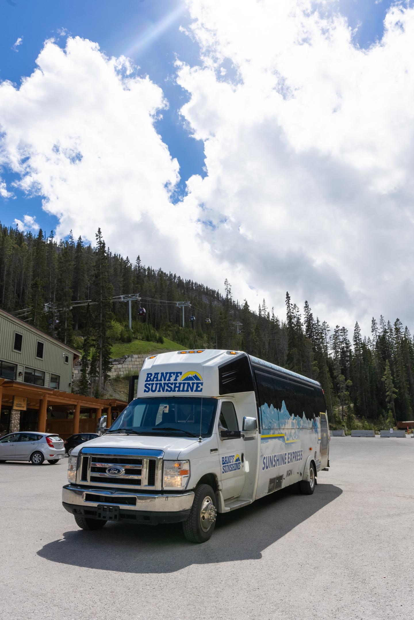 White shuttle bus parked beside a forested mountain road under cloudy sky.