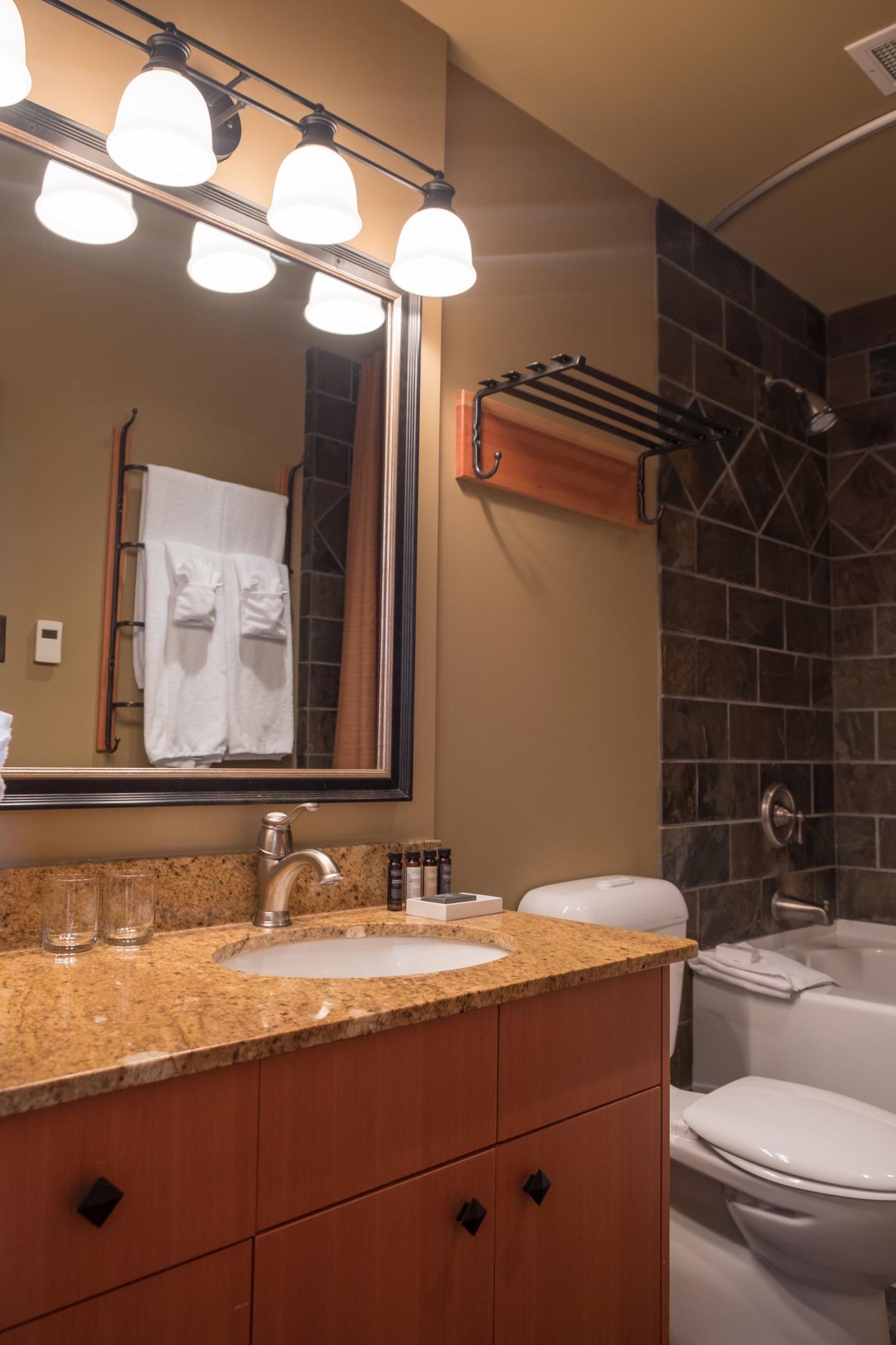 Warm bathroom with wooden vanity, double sinks, black tile shower, and toilet.