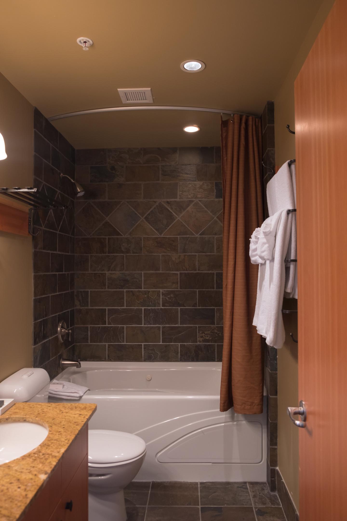 Small bathroom with a bathtub, toilet, wooden vanity, and dark tiled shower walls.