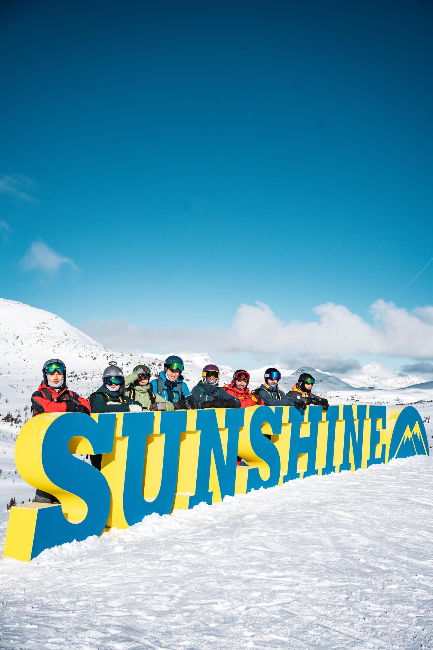 People in winter gear behind a large "Sunshine" sign in snowy mountains.