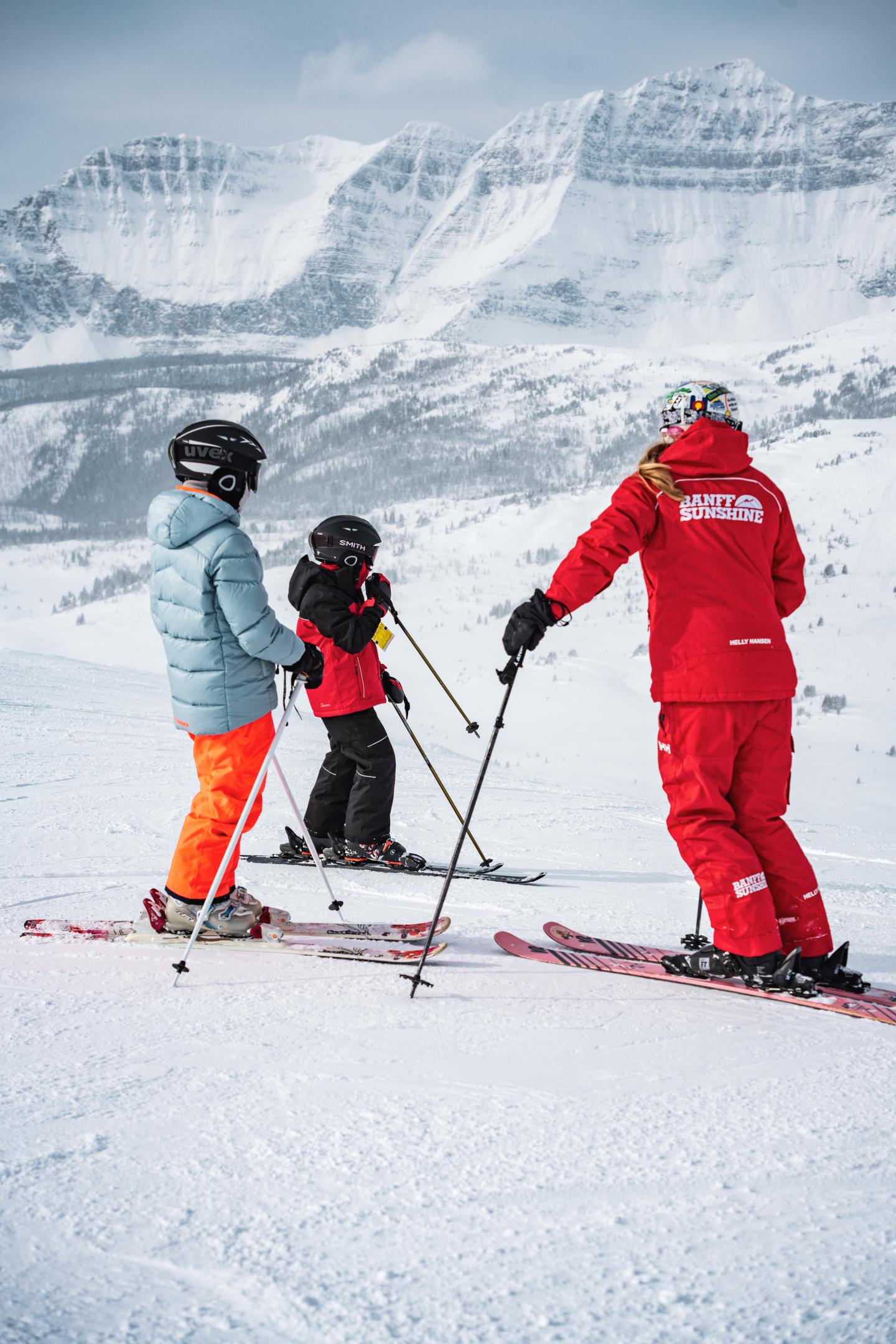 Skiers in colorful gear on snowy mountain slope, with snowy peaks in the background.