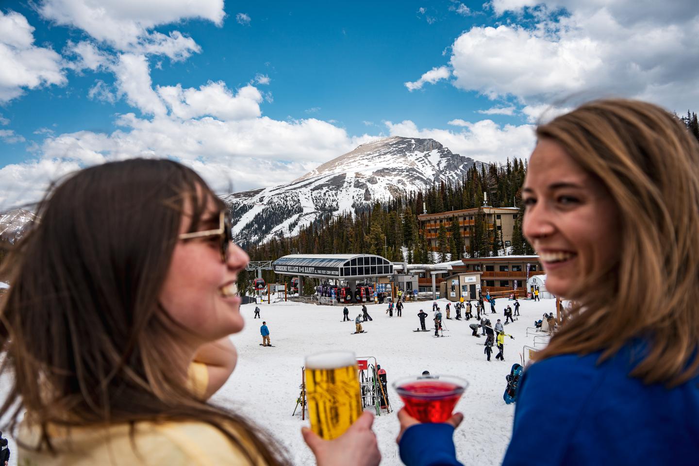 Two women smiling outdoors with ski slopes and snowy mountains behind them.