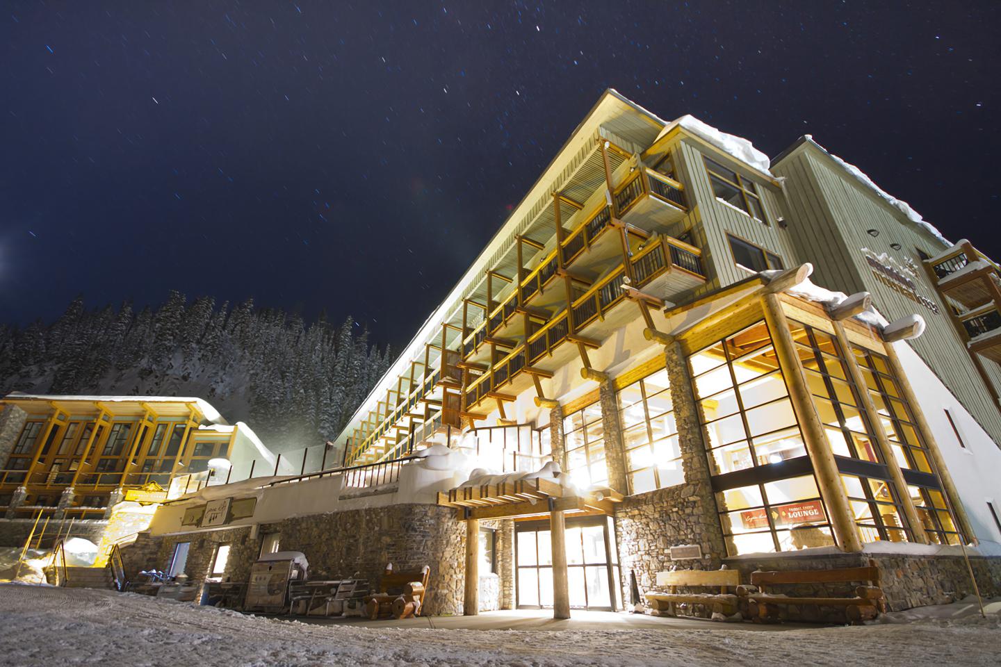 Night view of a lit, modern mountain lodge under a starry sky.